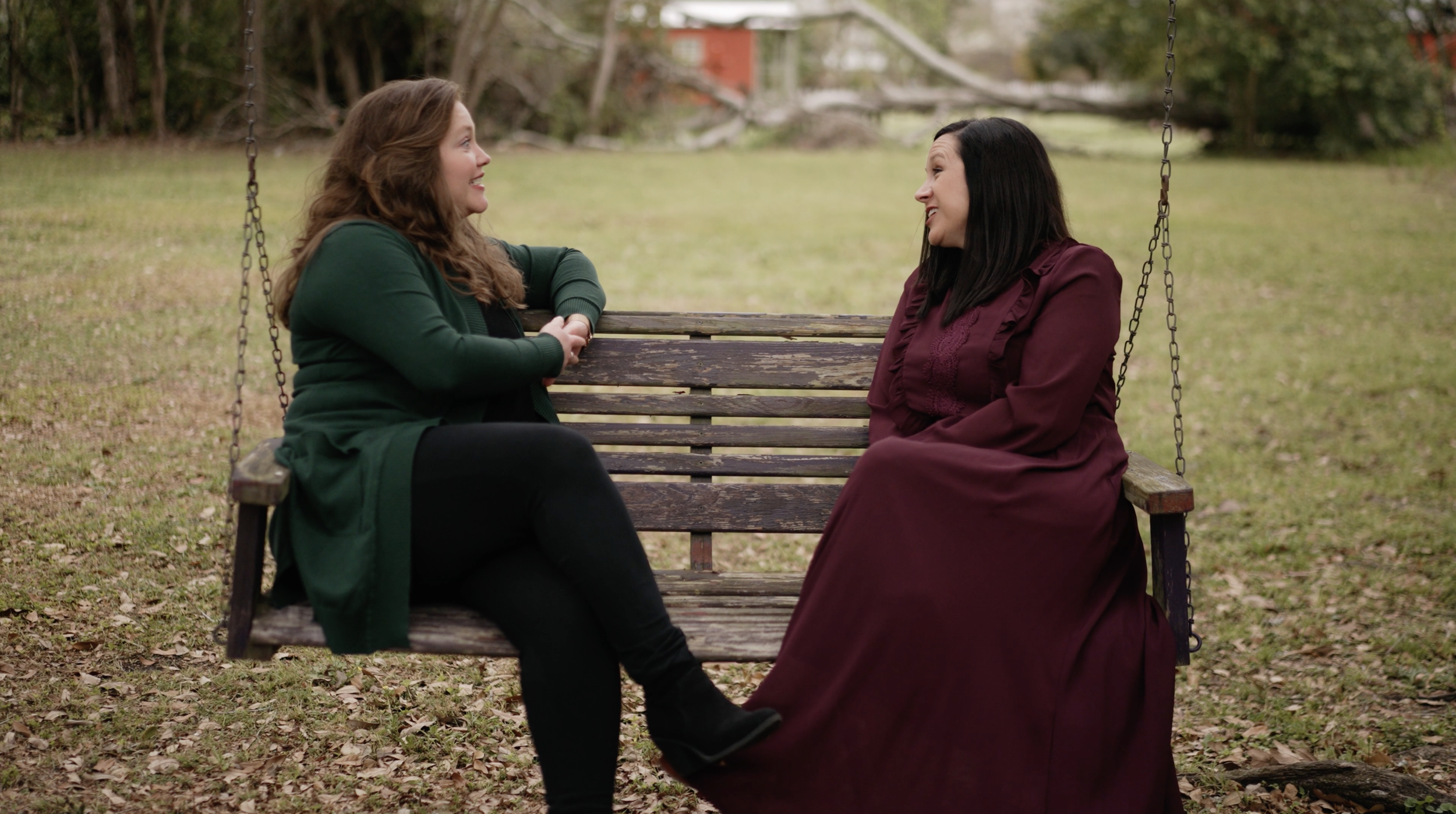 Two women sitting on a wooden swing bench outdoors, engaging in a conversation, with trees and grass in the background.