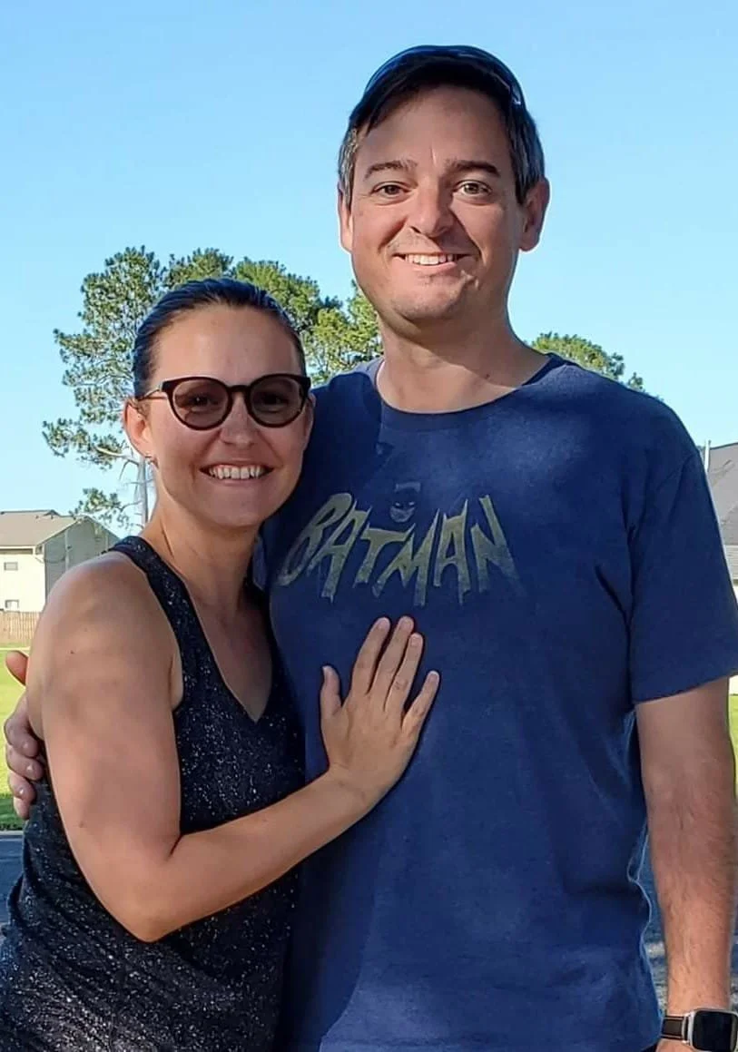 A smiling couple standing outside, embracing each other. The woman is wearing glasses and a sparkly sleeveless top, and the man is wearing a blue Batman t-shirt. The background features trees and houses under a clear blue sky.
