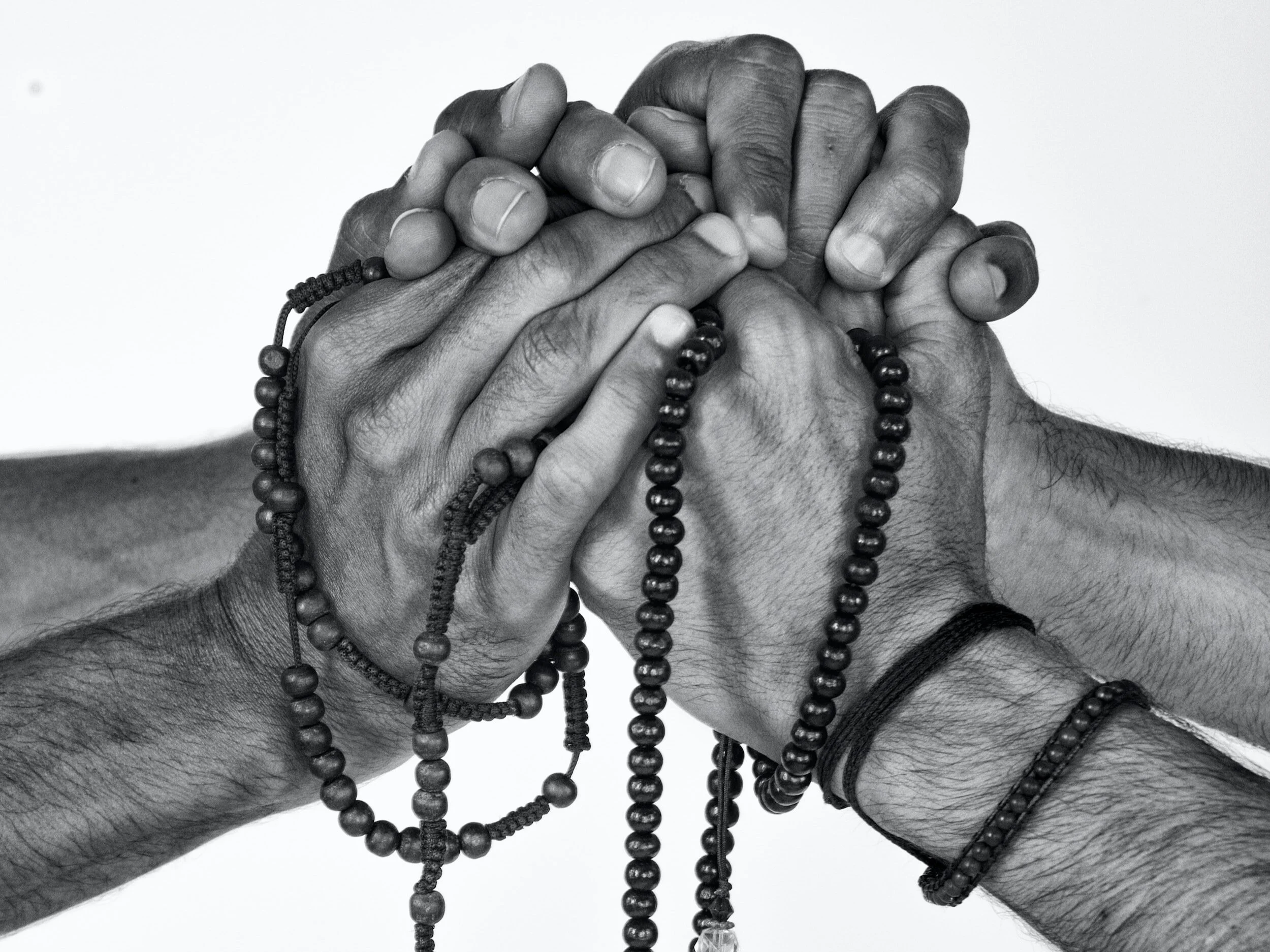 Two pairs of hands clasped together, holding prayer beads, in black and white.