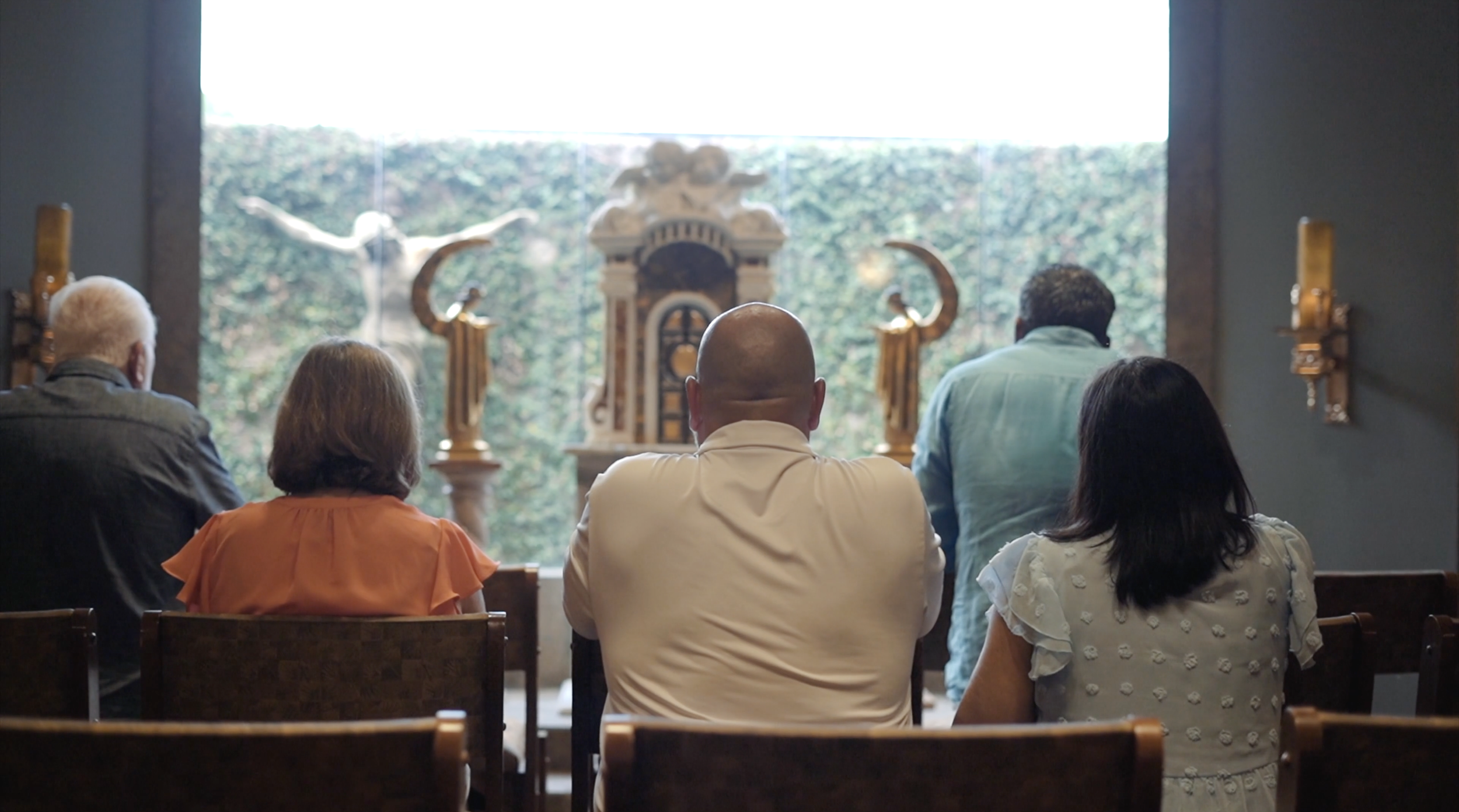 Five individuals seated in a chapel, facing an altar with religious statues and a window showing greenery outside.