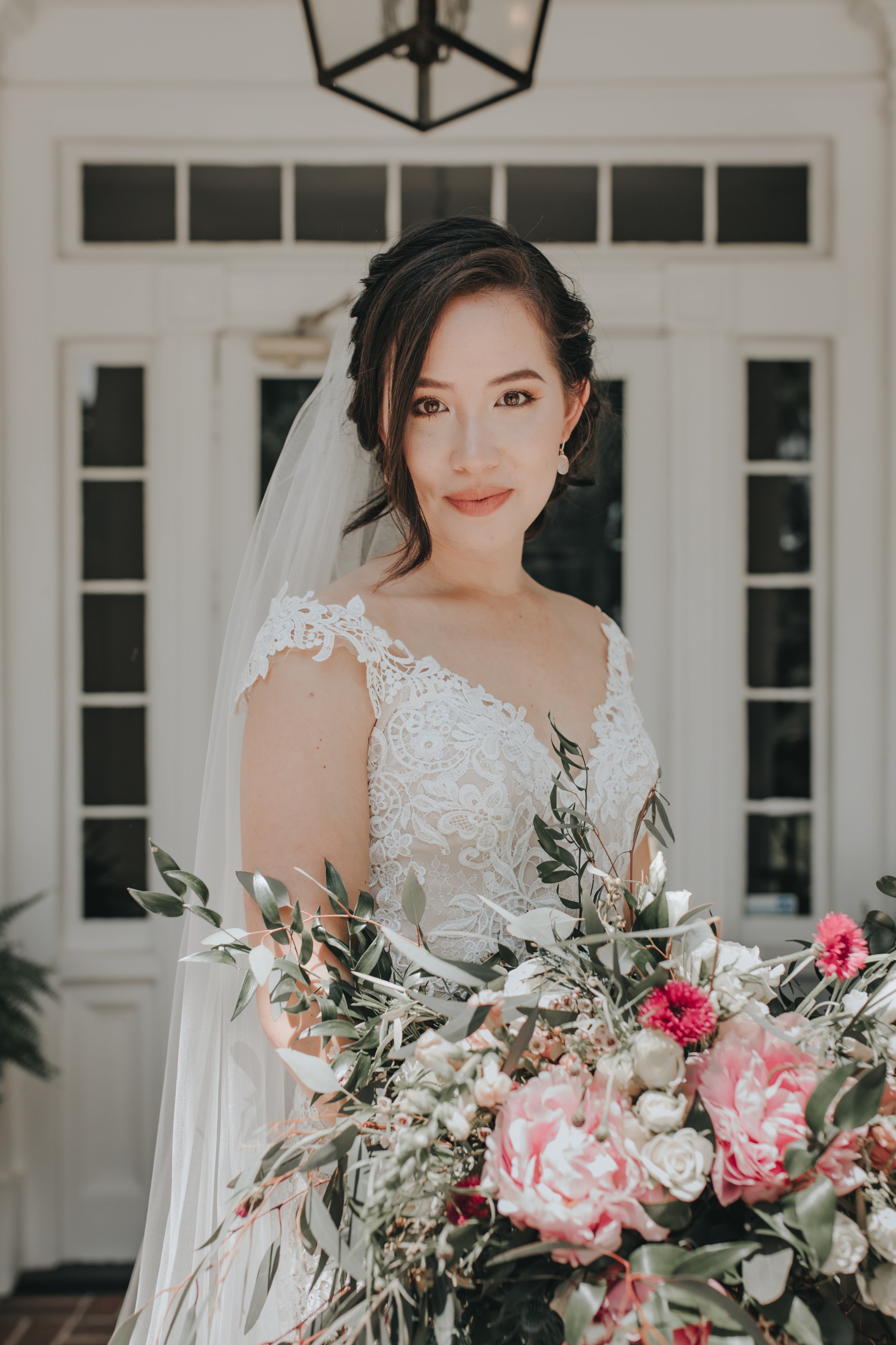 A bride in a lace wedding dress holding a large bouquet of pink and white flowers, standing in front of a door with a black lantern overhead.