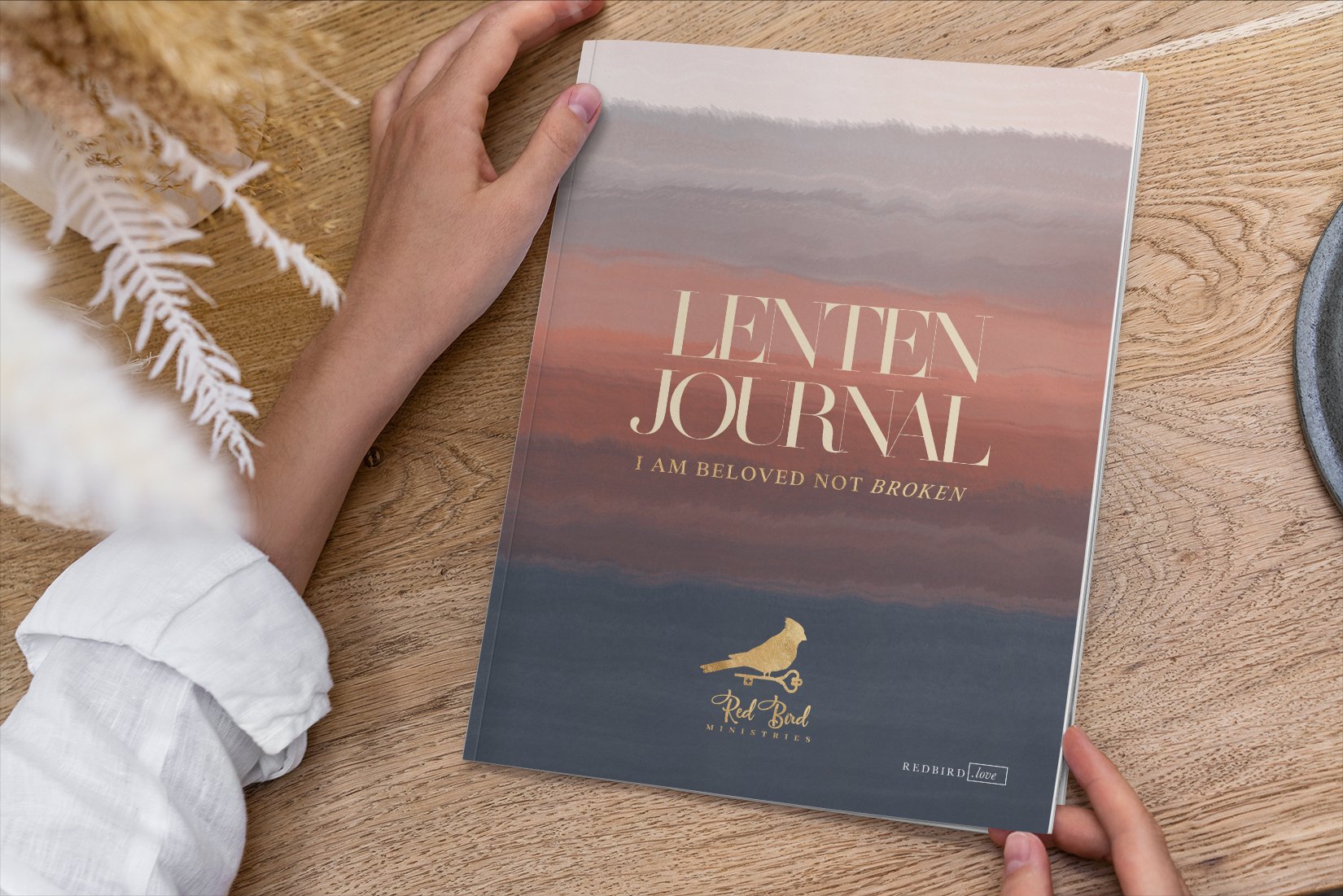 A person holding a pink and blue sunset-themed Lenten journal with the words "Lenten Journal I Am Beloved Not Broken" on the cover, placed on a wooden table.