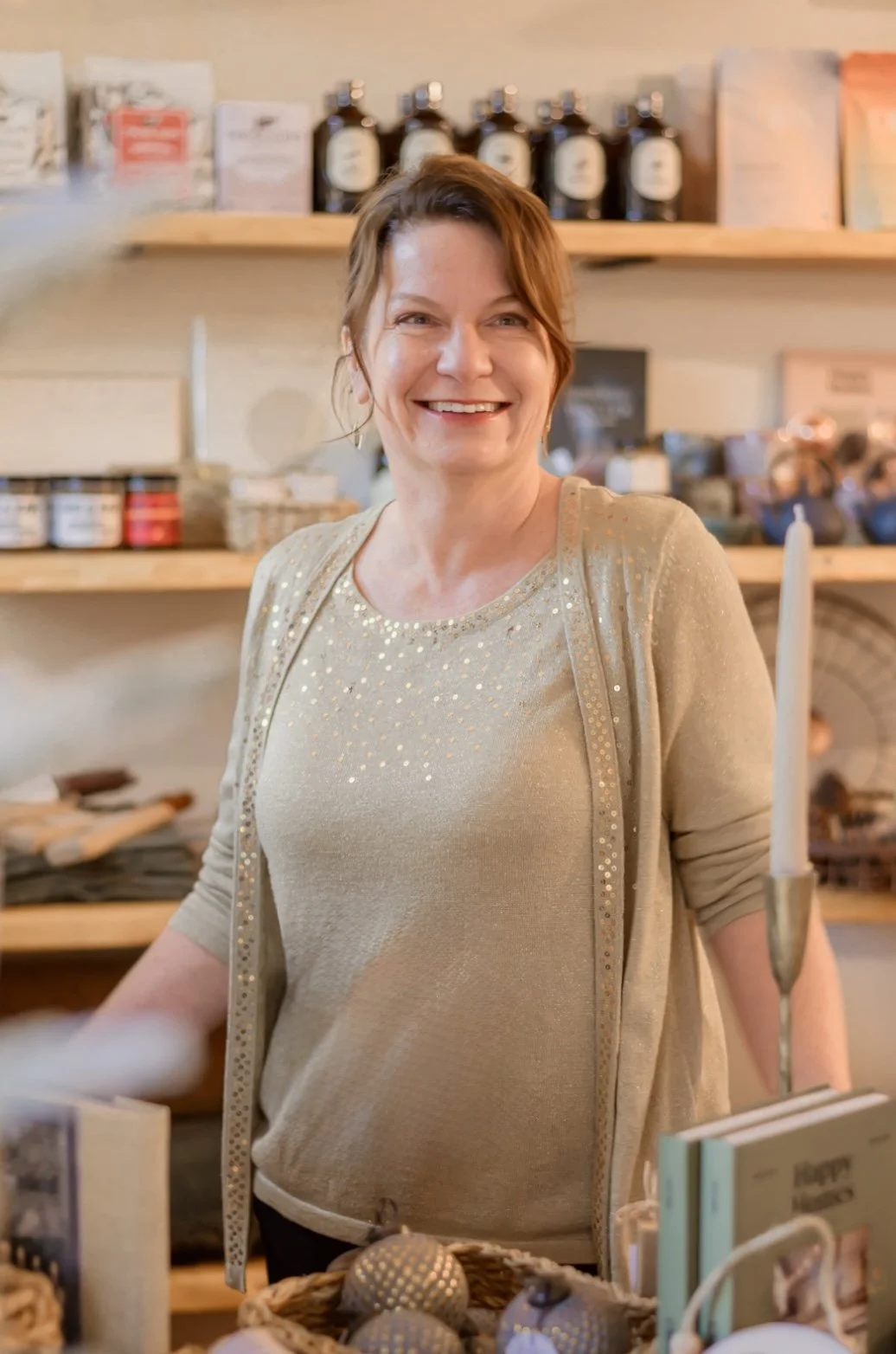 A woman with short brown hair, wearing a beige sweater with glittering gold dots, smiling in a store with wooden shelves filled with various products behind her.