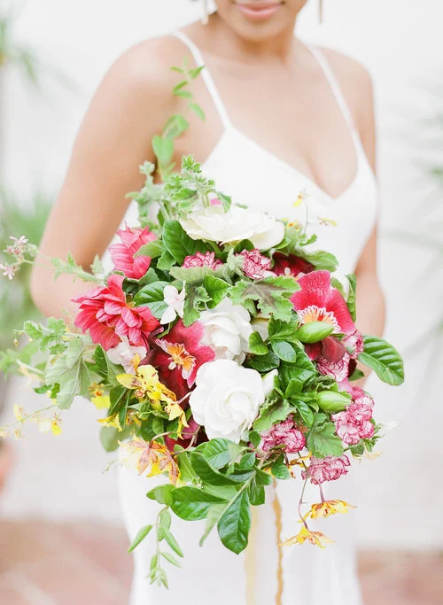 Woman in white dress holding a large colorful bouquet of pink, white, and yellow flowers with green leaves.