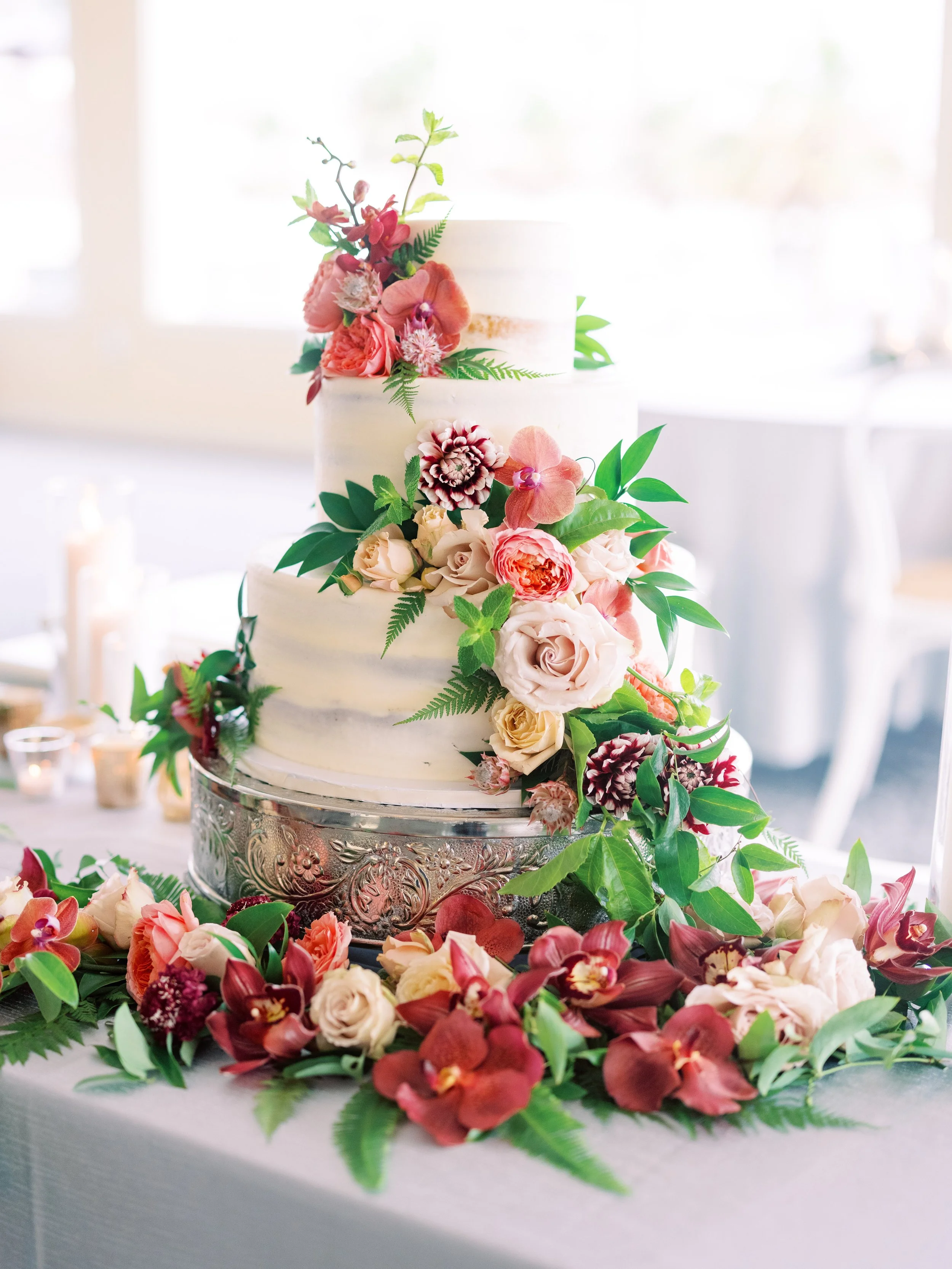 A three-tiered wedding cake decorated with pink, white, and purple flowers and green leaves, surrounded by more flowers on a table.
