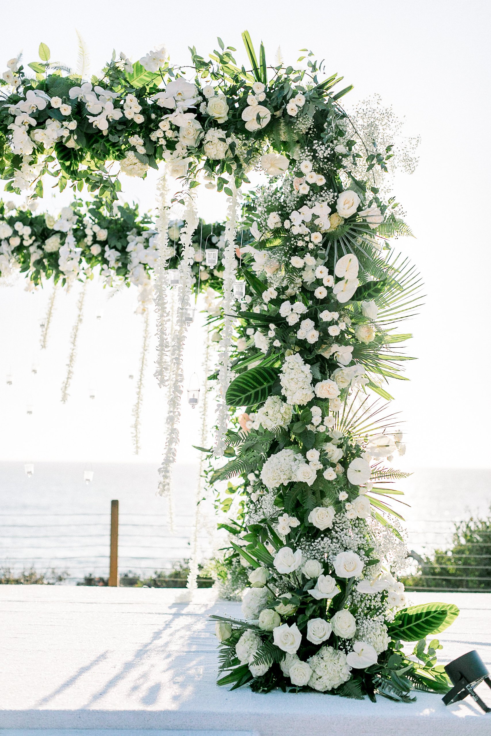 White floral wedding arch with roses, orchids, and greenery overlooking the ocean.