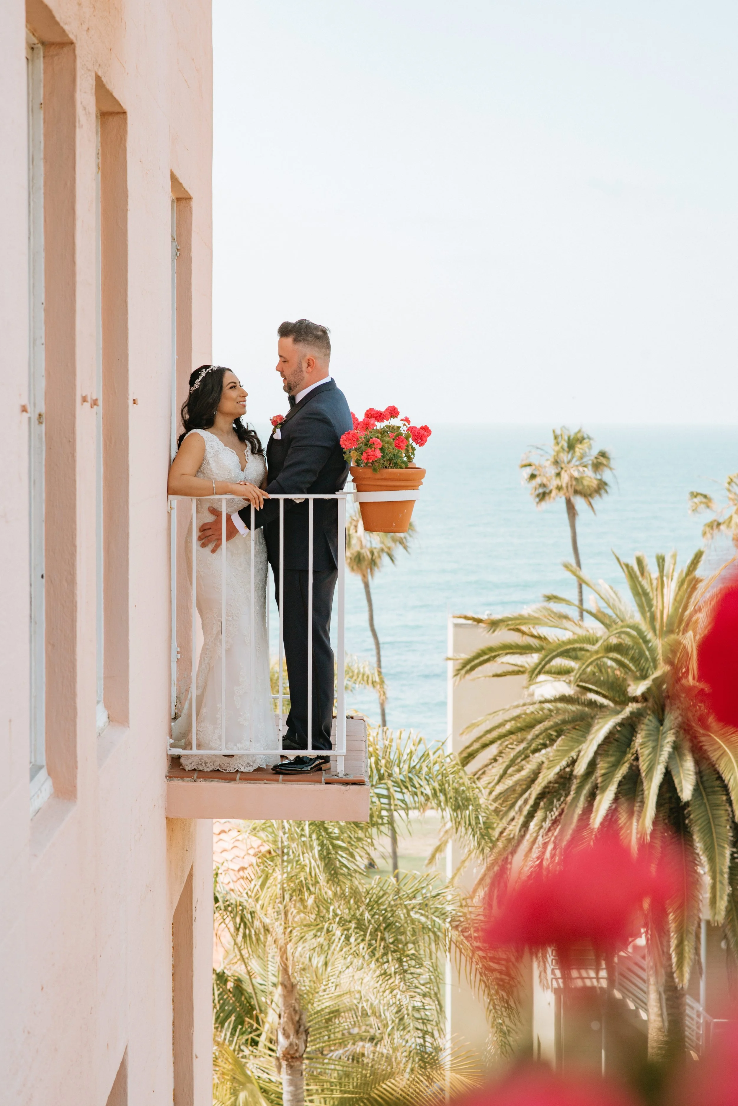 A bride and groom stand on a pink balcony overlooking the ocean and palm trees, sharing a moment during their wedding.