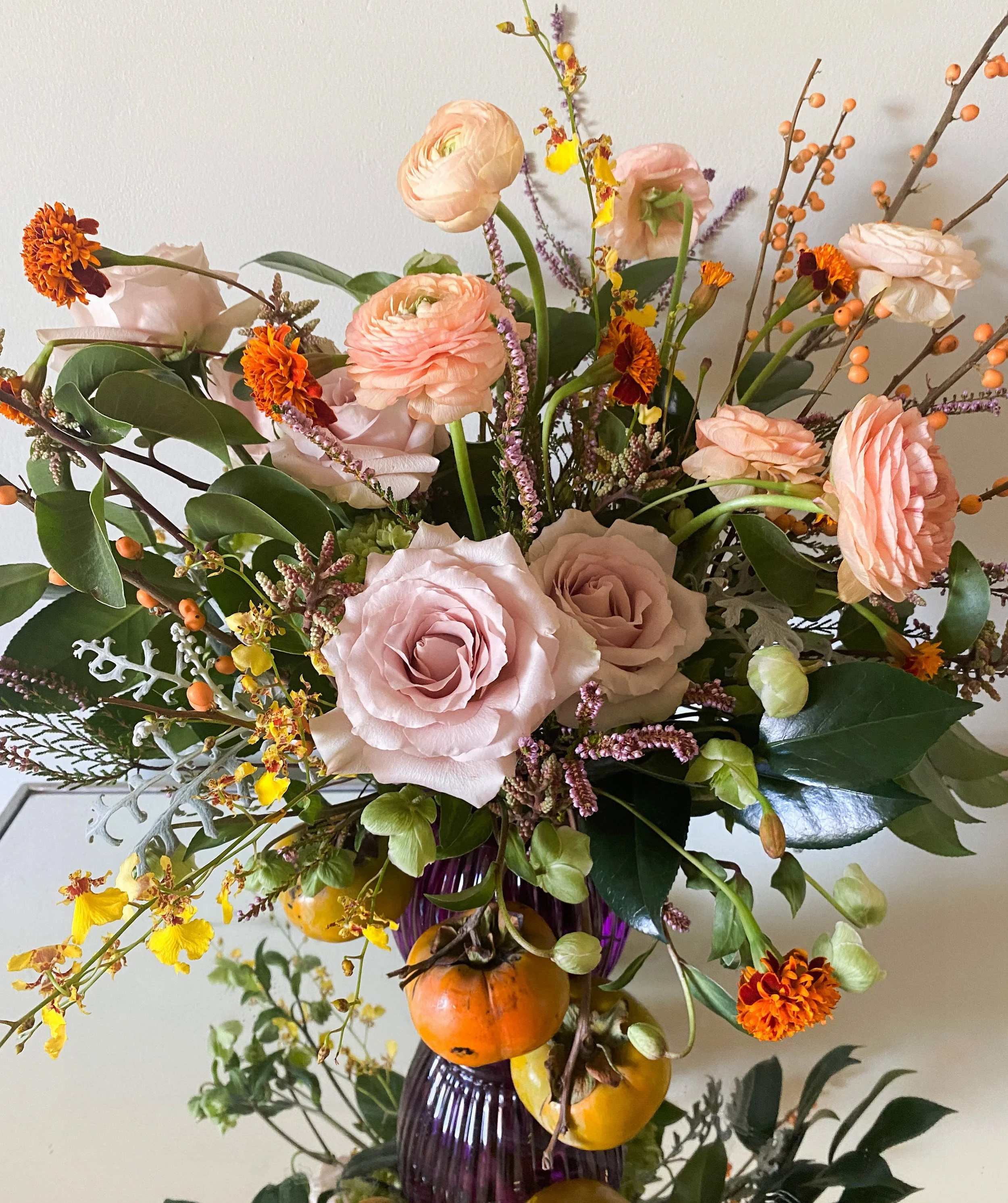 A floral arrangement in a purple vase with pink roses, peach ranunculus, orange marigolds, yellow flowers, and foliage, including persimmons hanging below.