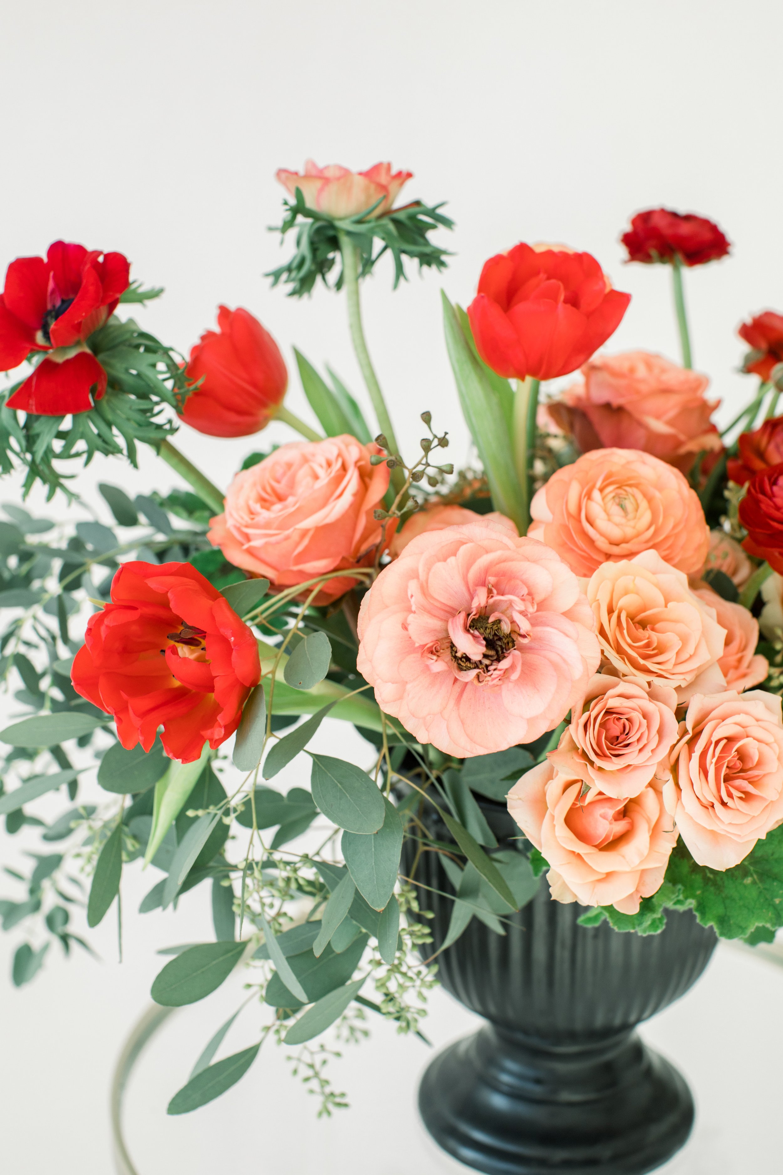 A black vase filled with pink, peach, and red flowers and green foliage against a plain white background.