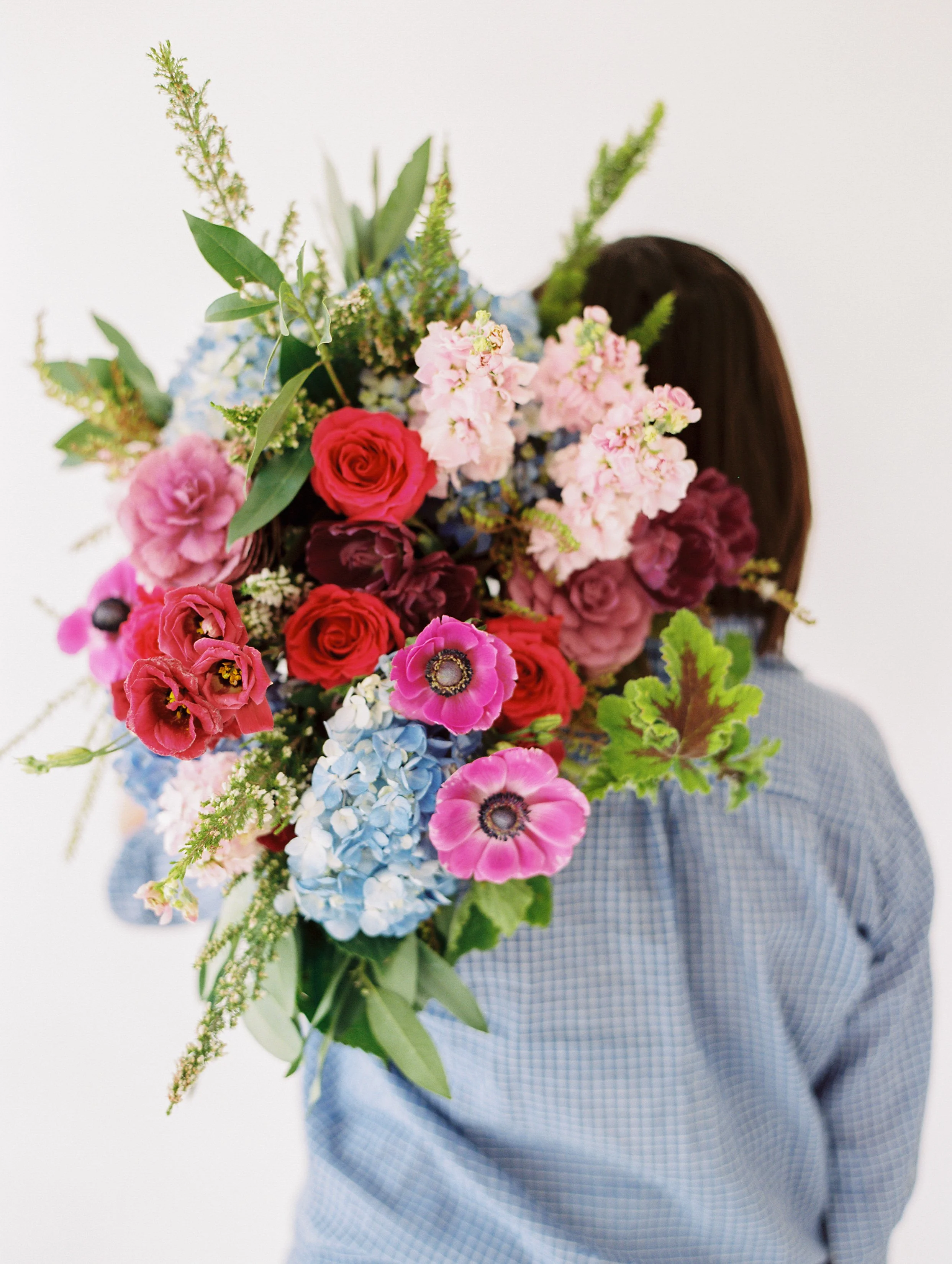 Person holding a large, colorful bouquet of pink, red, purple, and blue flowers in front of their face, wearing a blue gingham shirt.