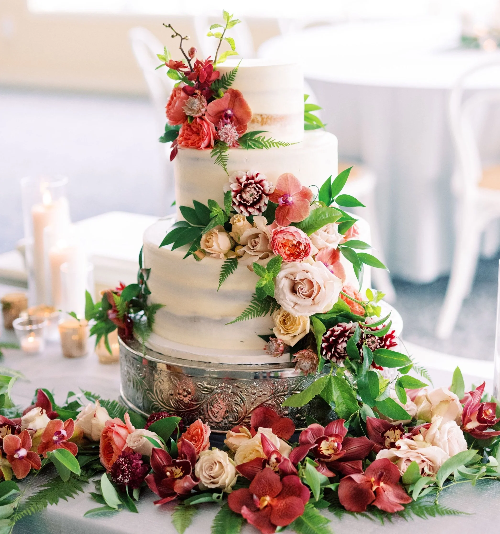 A tiered wedding cake decorated with pink, peach, and cream flowers, surrounded by an arrangement of similar flowers and green foliage on a table.
