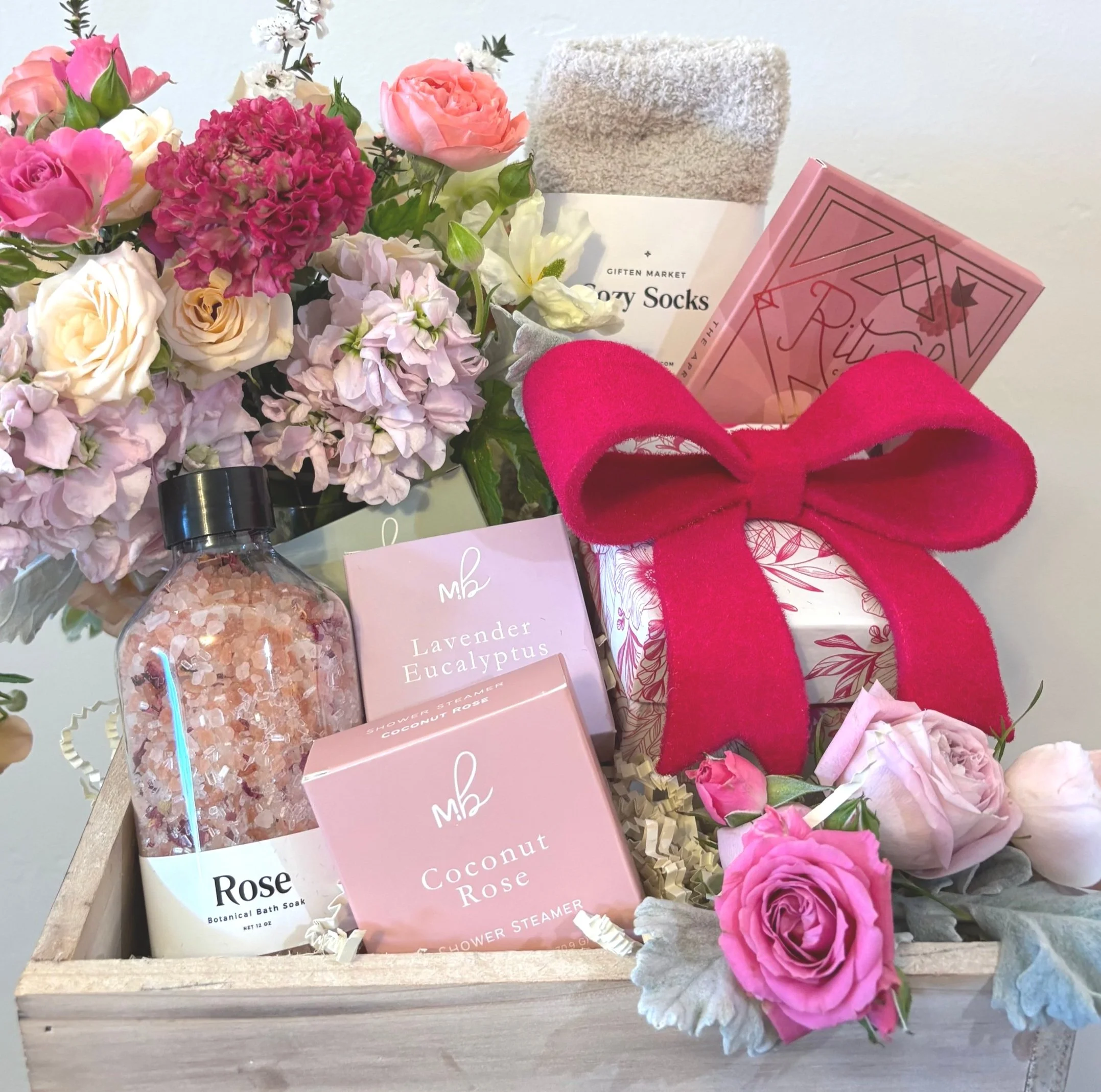 Gift basket with pink roses, peonies, and lavender eucalyptus flowers, containing bath soaks, bath salts, a pink bow, and other self-care items.
