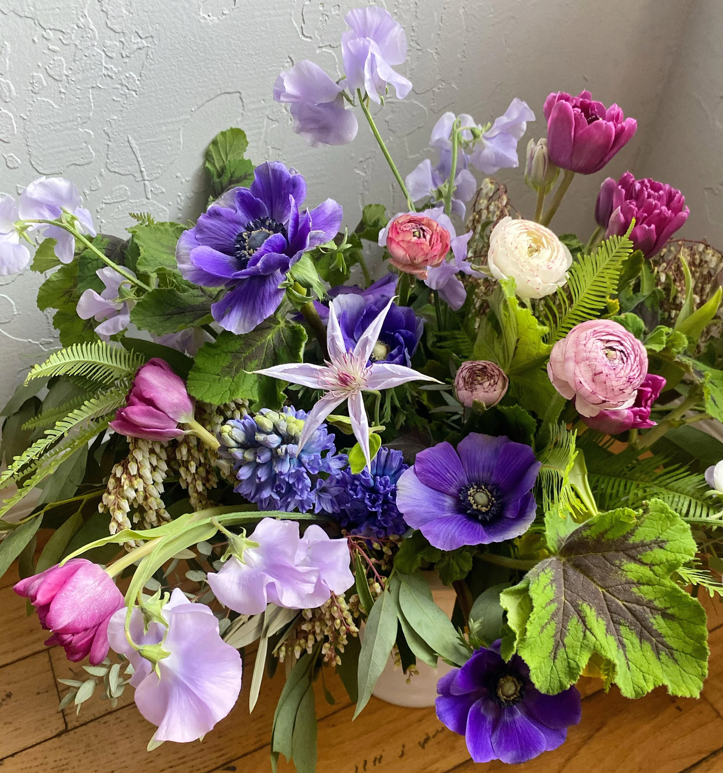 A colorful arrangement of various purple, pink, and white flowers with green leaves in a white vase on a wooden surface.