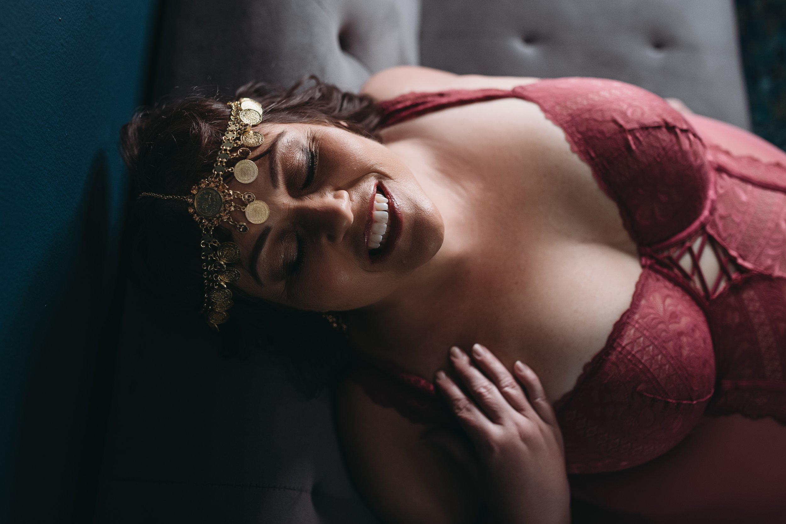 A woman over 40 with dark hair wearing a gold coin headpiece lying on a dark couch, smiling and wearing a pink lace dress.