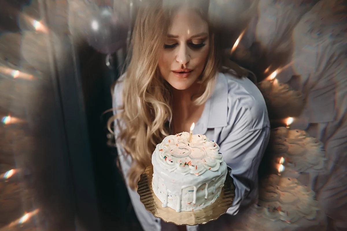 A woman with long blonde hair holding a whipped cream decorated birthday cake with a single lit candle celebrating her birthday
