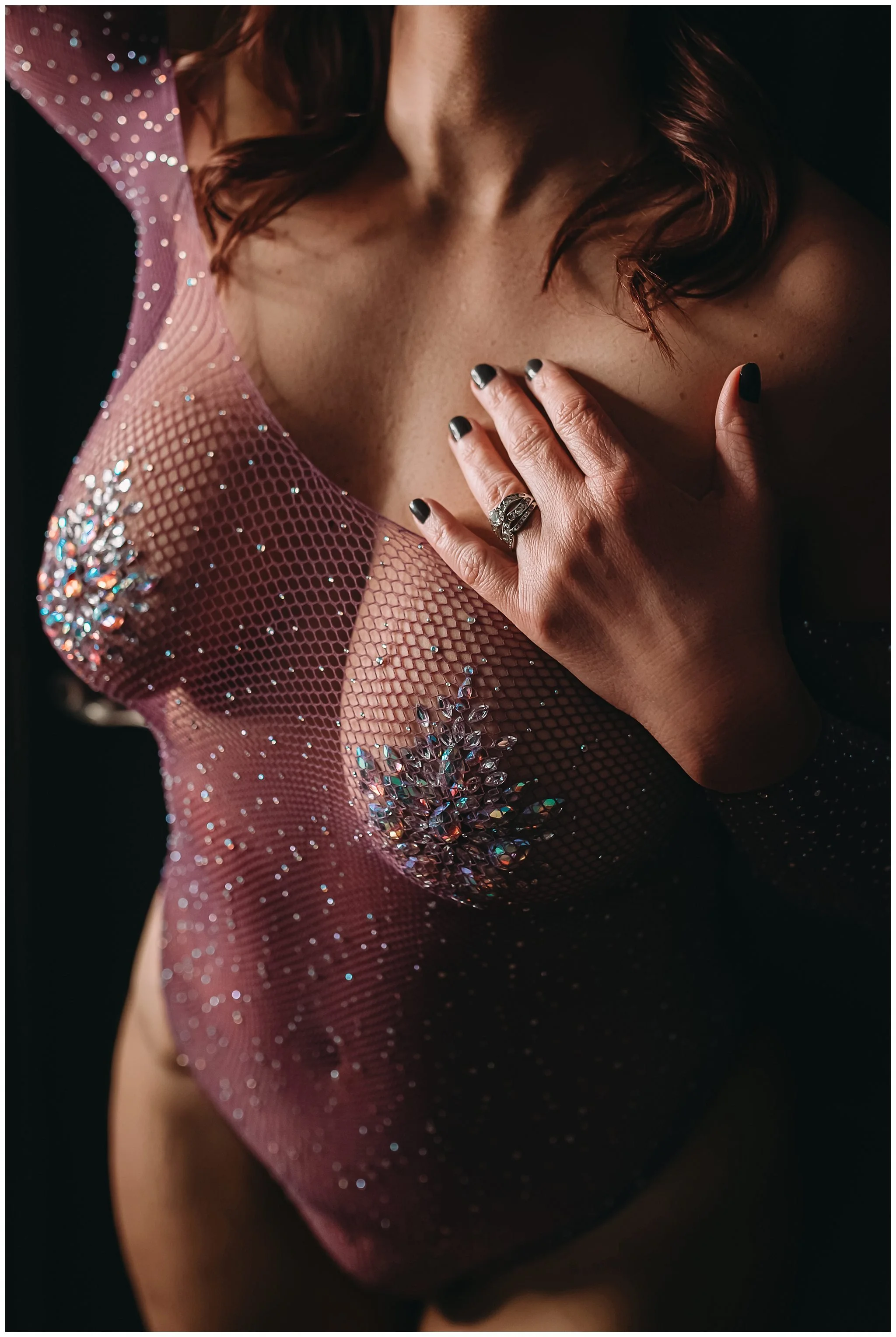Boudoir client poses with hand on chest as she poses in purple bodysuit with jewel pasties during photography session