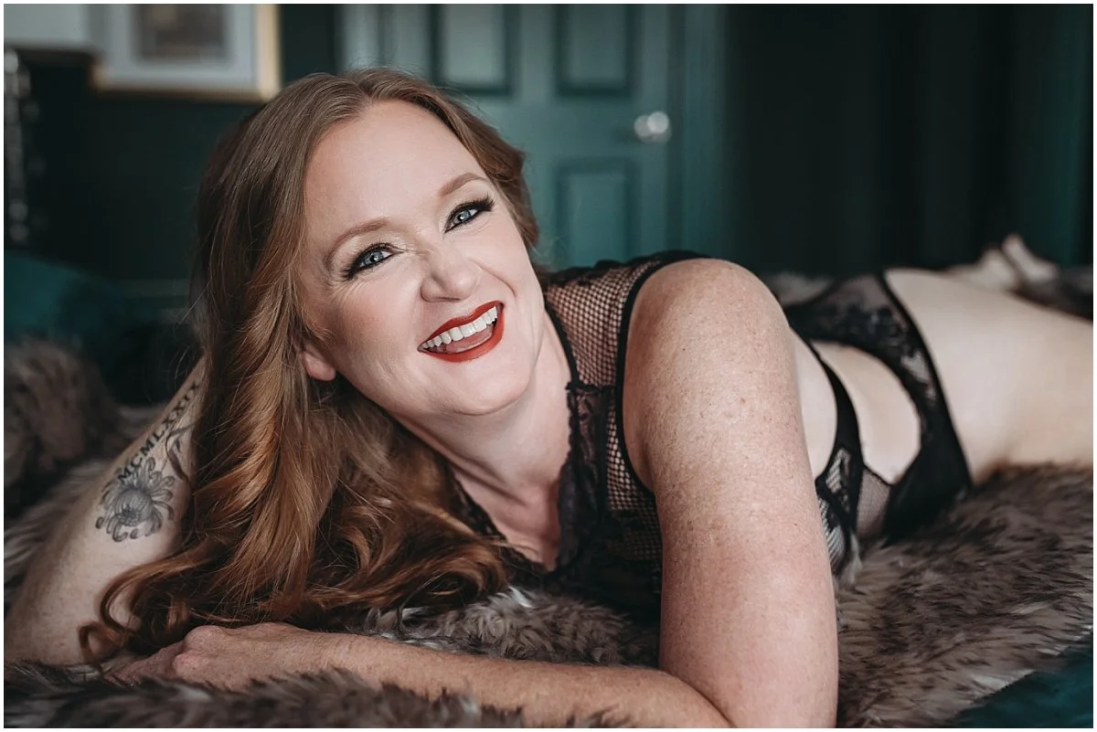 Boudoir photography for women over 40 featuring a redhead smiling on a bed in a dark, intimate studio space