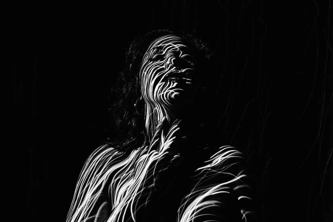 A black and white artistic portrait of a woman with light streaks across her face and body, creating a luminous pattern against a dark background.