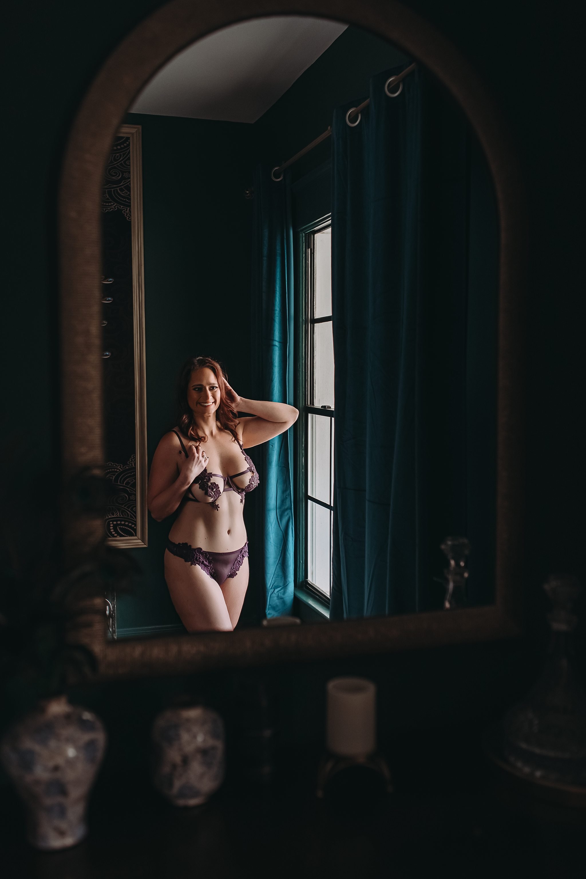 Client stands by window in dark and moody space in purple lingerie to celebrate weight loss journey and birthday