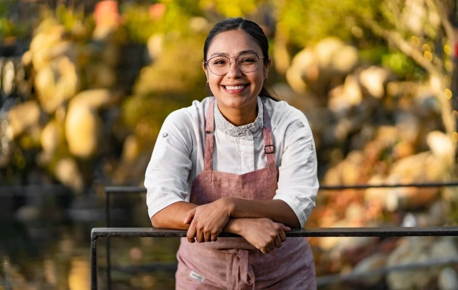 Mujer sonriendo, usando gafas y delantal, con un fondo de naturaleza, en un entorno al aire libre.