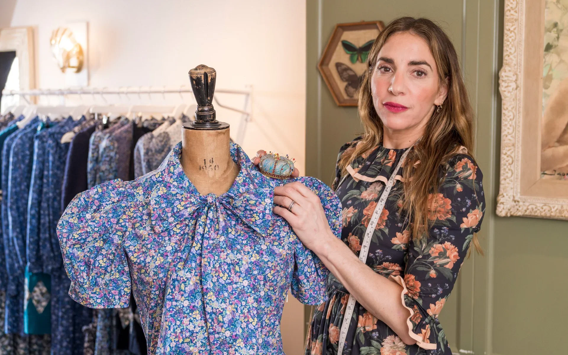 woman with long brown hair, pins needles into a dress makers dummy that has a floral dress on it.