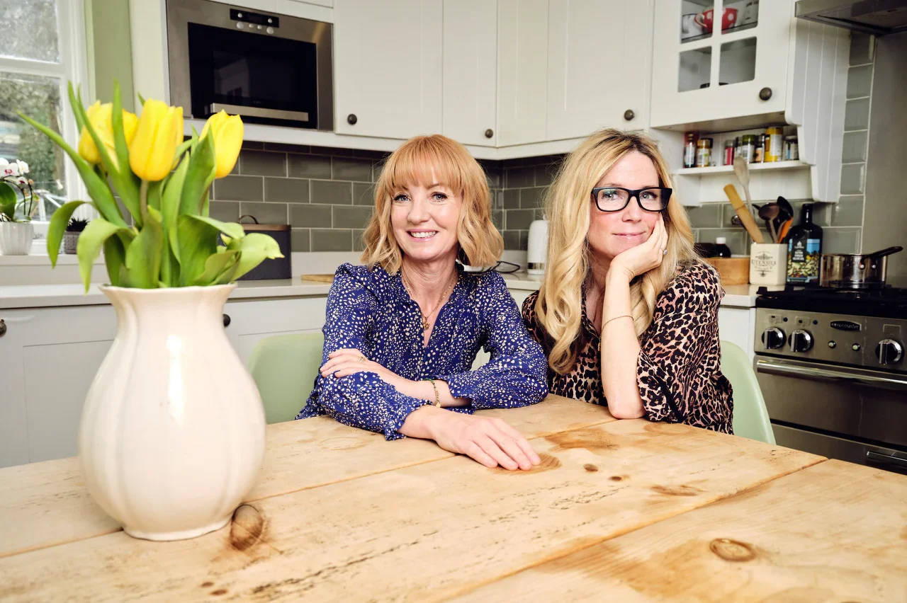 two women with blonde hair sit at a wooden table in a kitchen, there are tulips in the foreground.