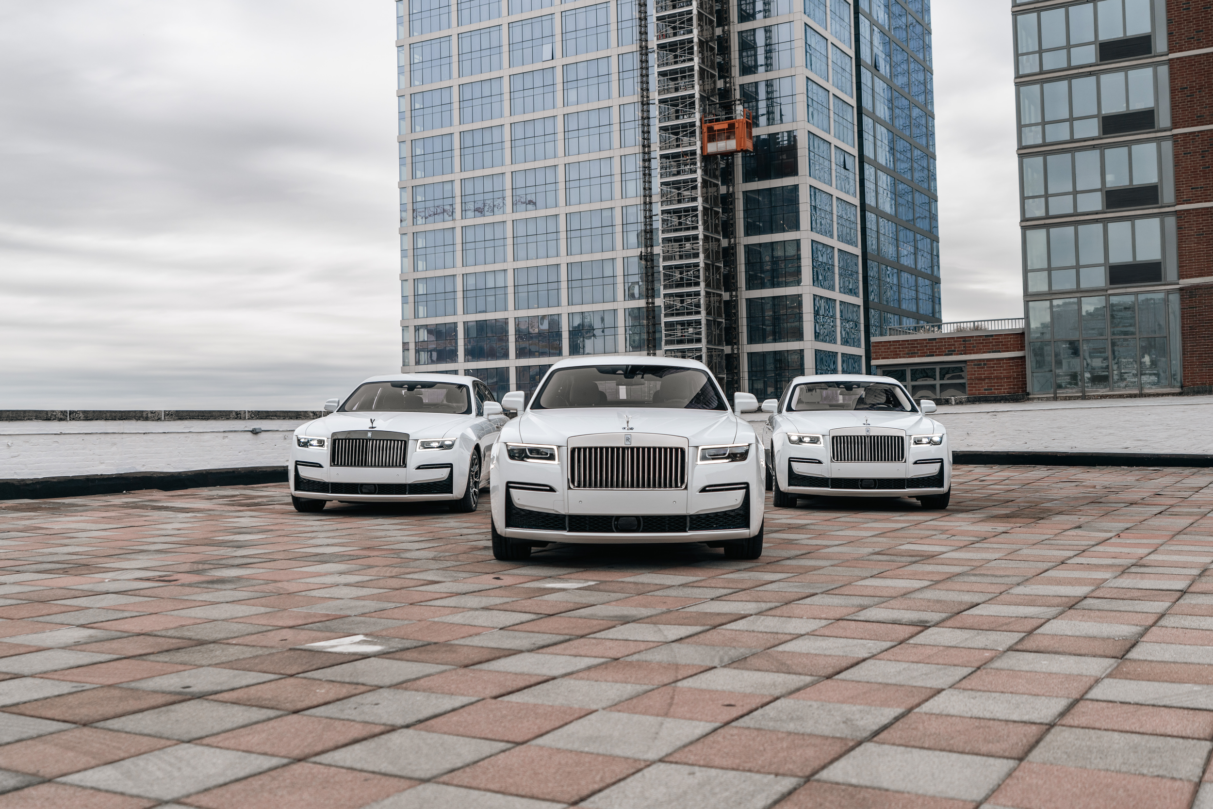 Three white luxury cars parked on a brick rooftop with a glass skyscraper under construction in the background, cloudy sky overhead.