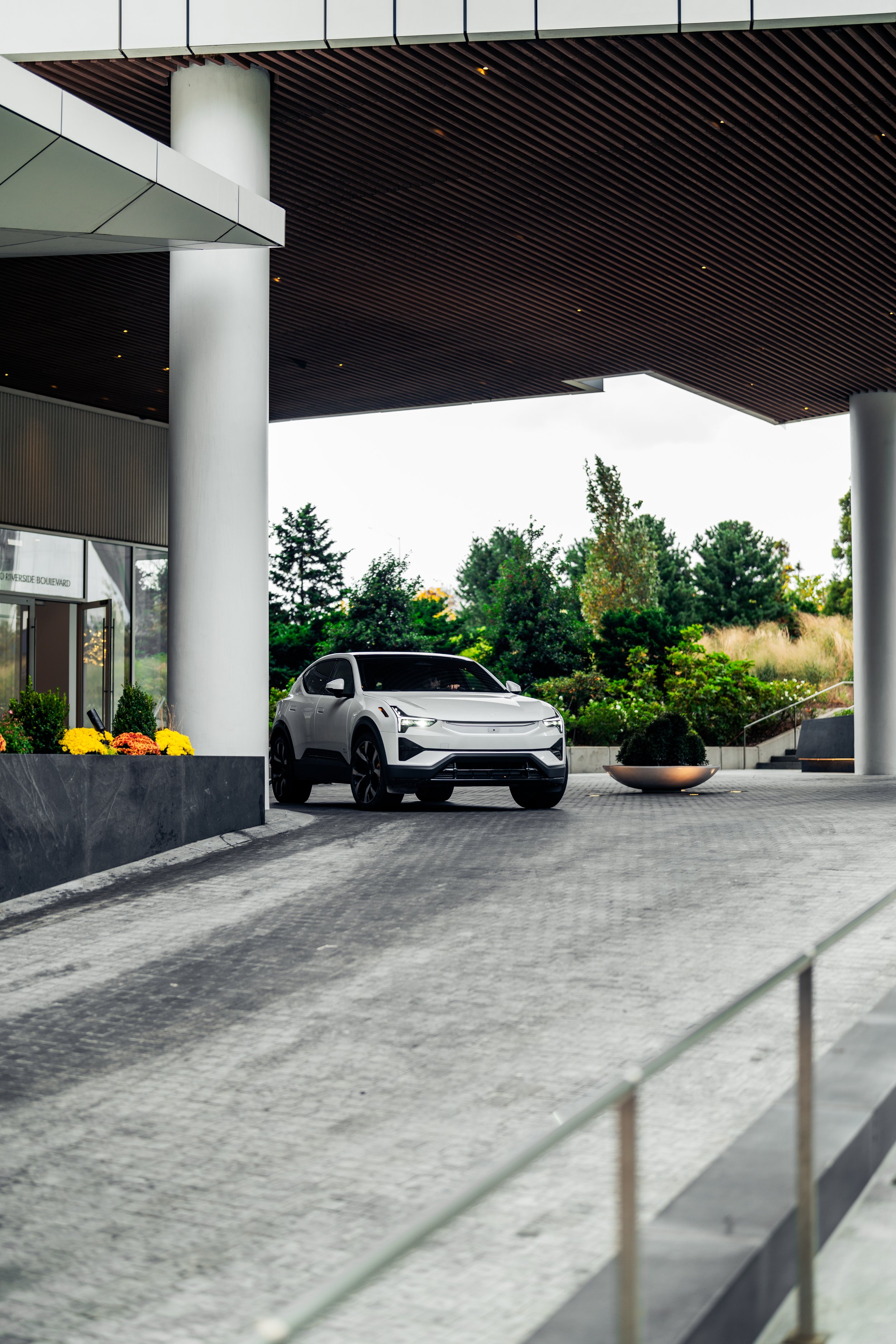 A modern building with large white columns and a wooden overhang. A Polestar 3 is parked underneath, surrounded by greenery and decorative flowers.