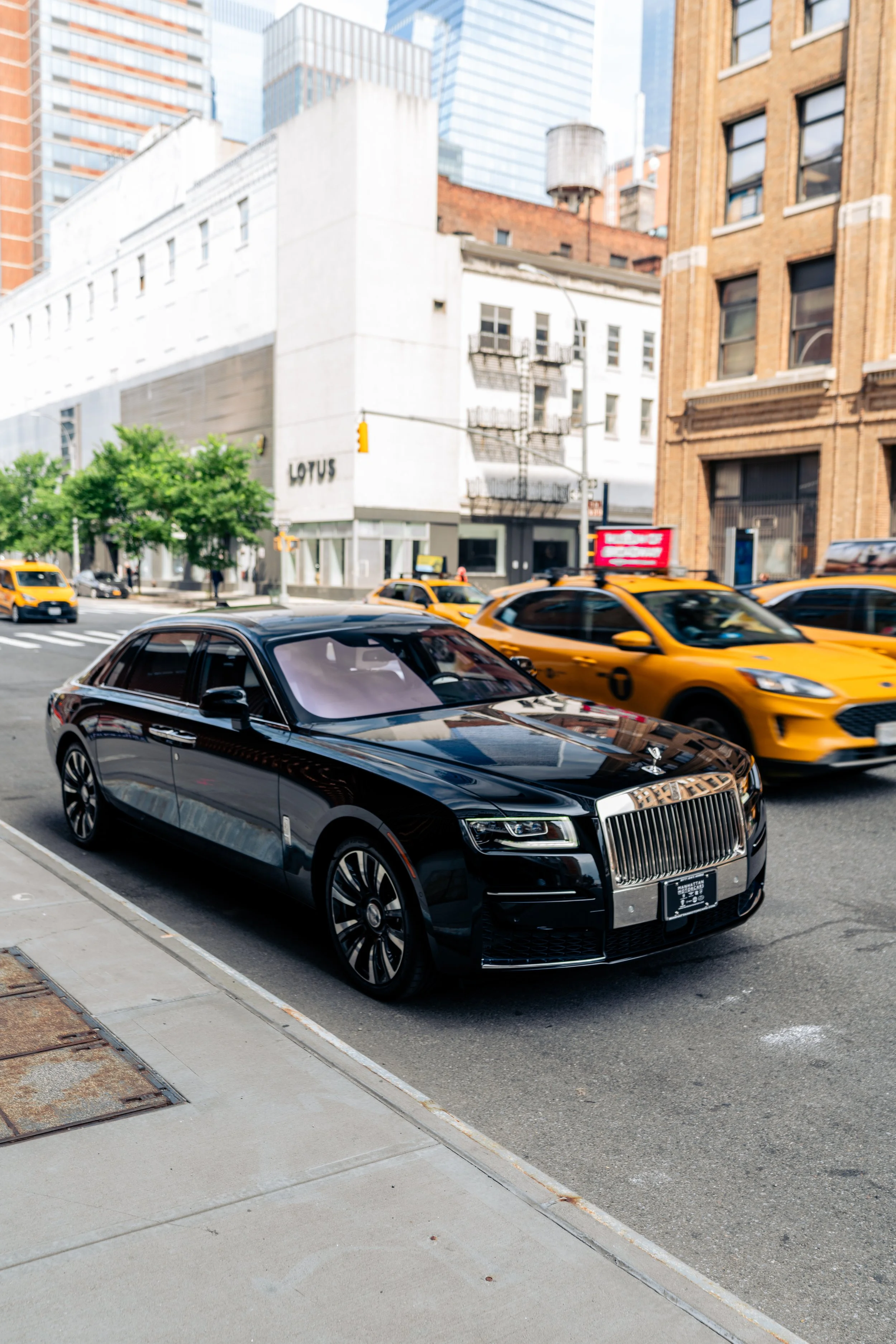 Black luxury car parked on city street with yellow taxis driving by, high-rise buildings in background, and a small tree on sidewalk.