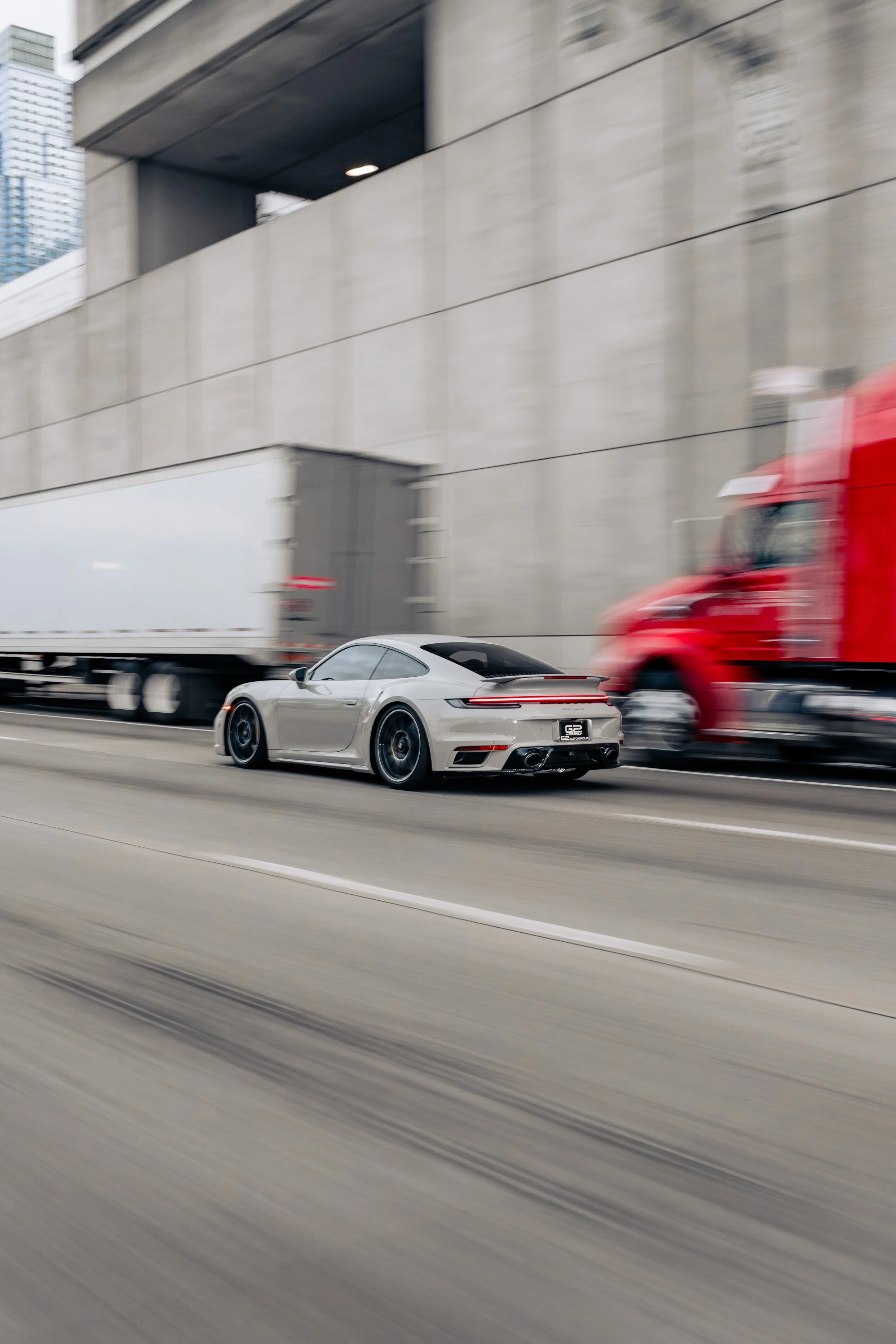 A silver sports car driving on a highway with a moving semi-truck and a red semi-truck passing by, against a backdrop of high-rise buildings and a concrete structure.