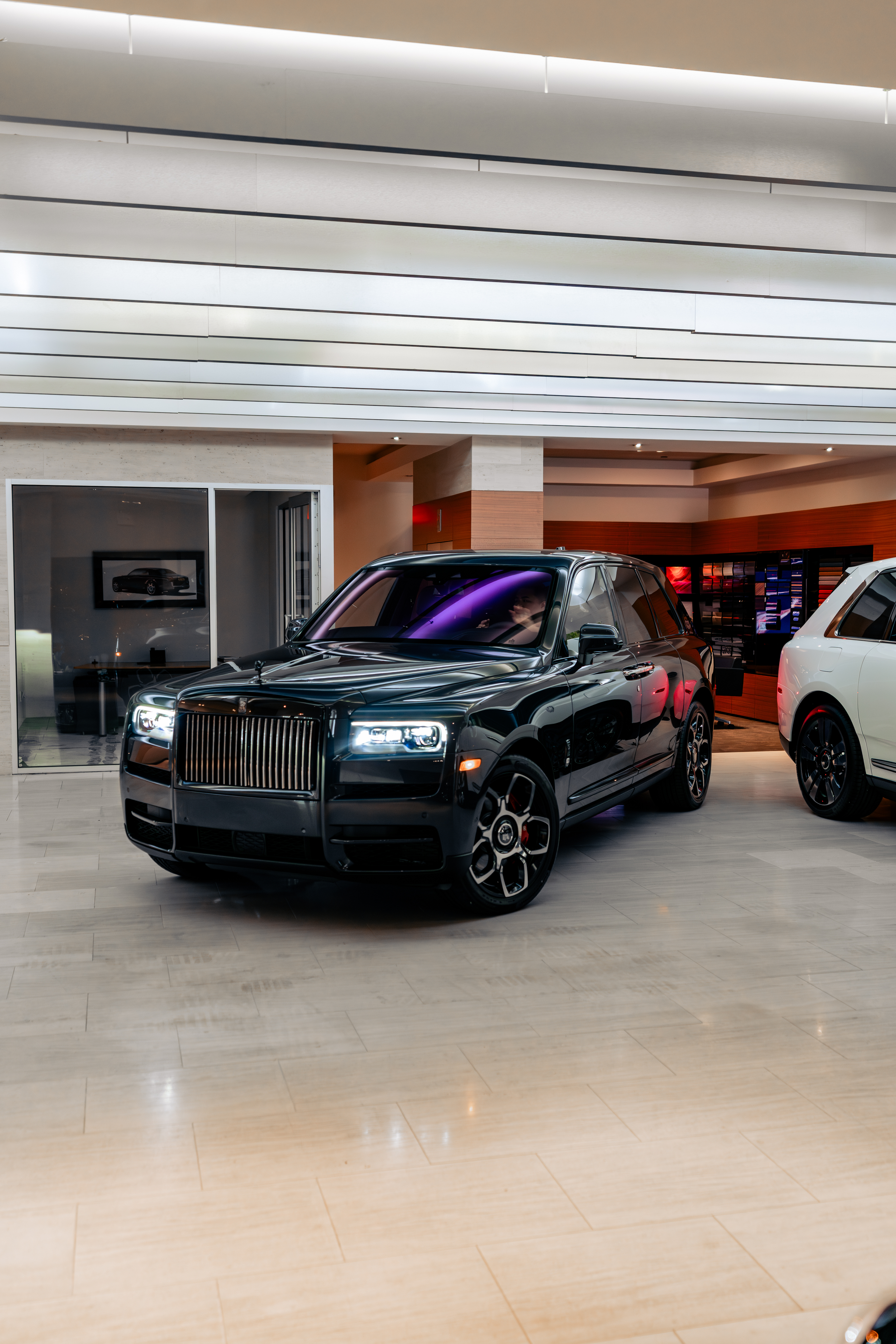Inside a luxury car dealership with a black Rolls-Royce SUV parked inside, showcasing a modern interior with beige and wooden accents and a promotional display on the wall.