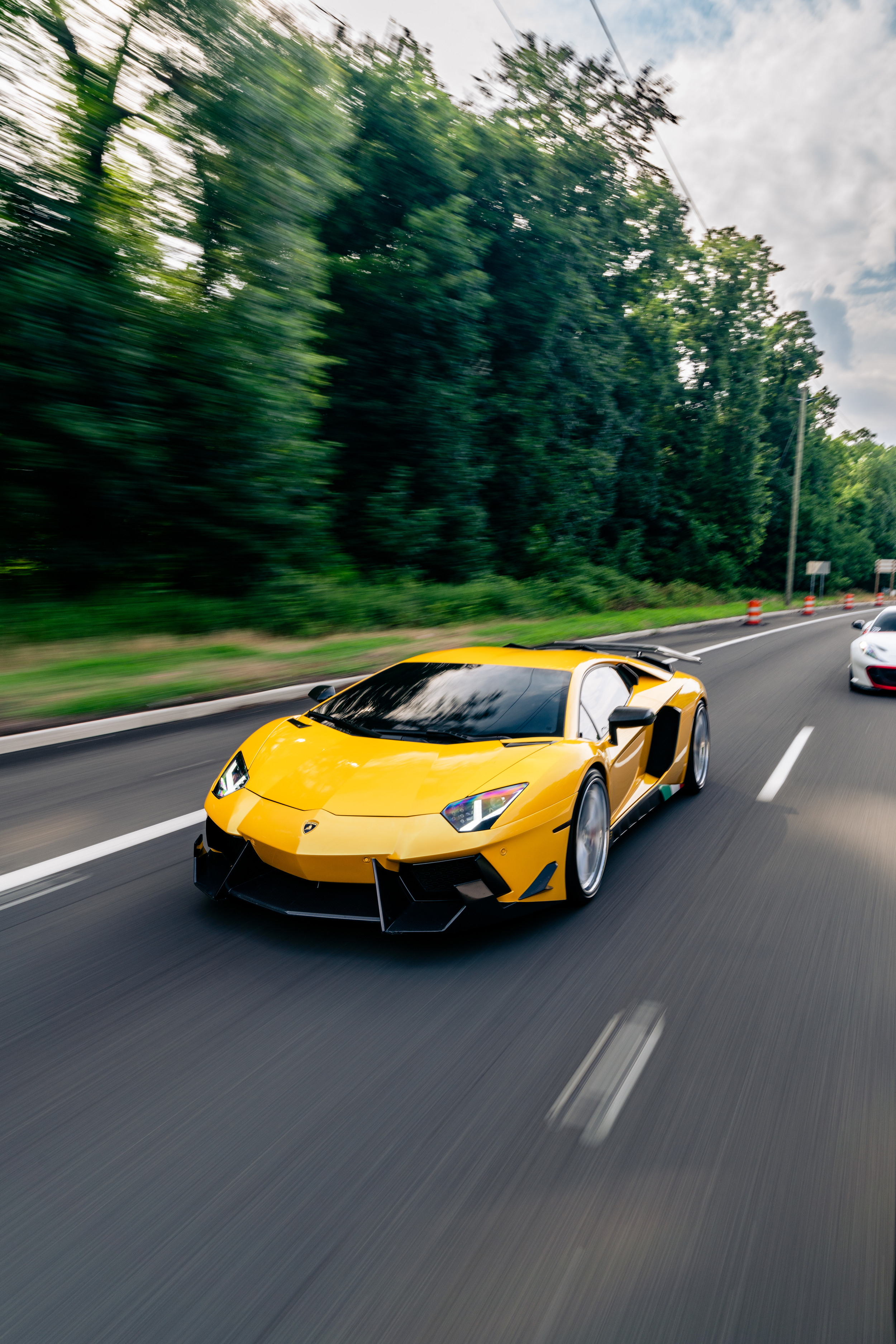 A yellow Lamborghini sports car driving on a highway with trees in the background and a partly cloudy sky.