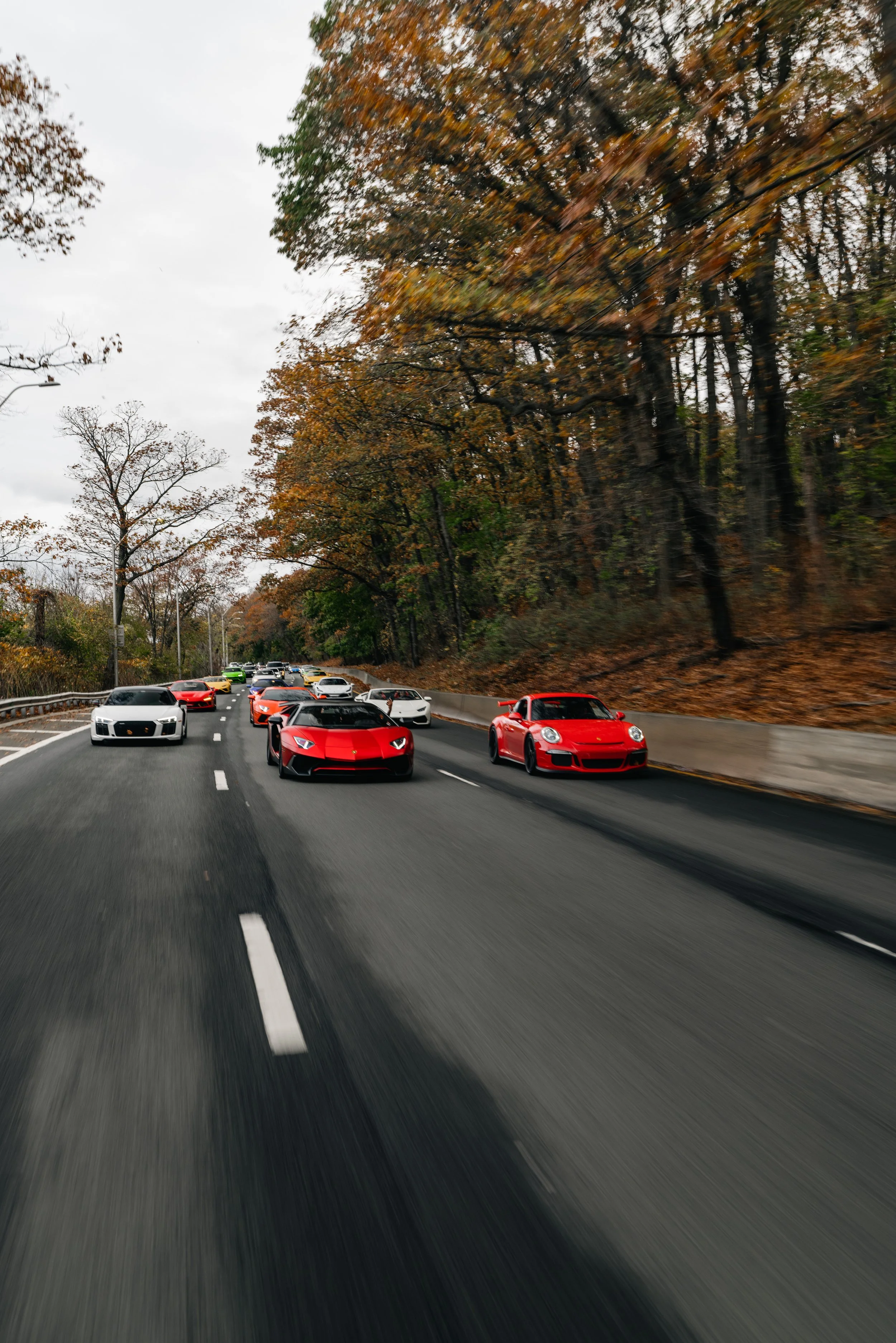 Red, orange, white, black, and yellow sports cars drive on a highway in a forested area during fall with trees having orange and brown leaves.
