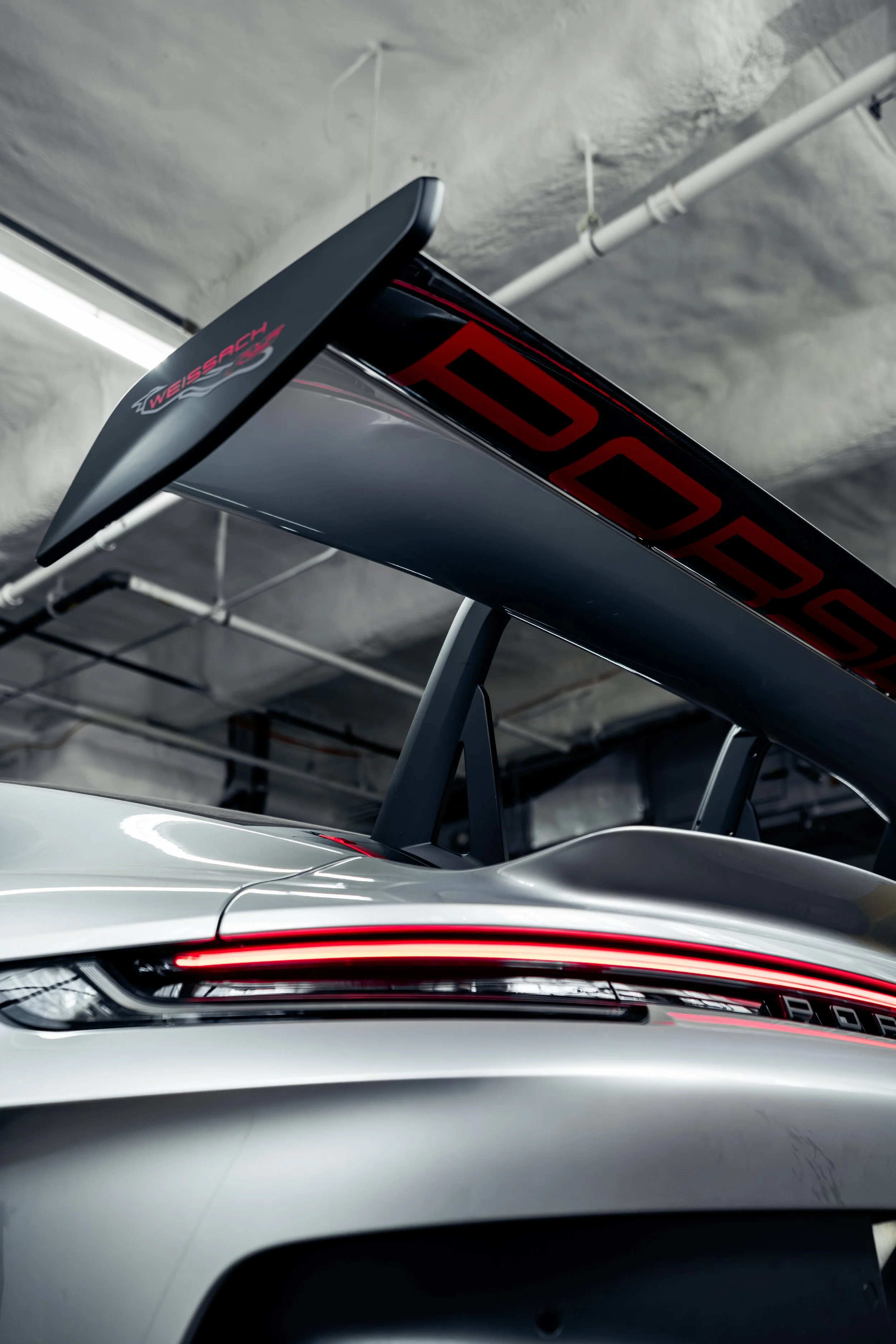 Close-up view of the rear wing of a racing car, featuring the 'Weissach' logo and aerodynamic design, set against a garage background.