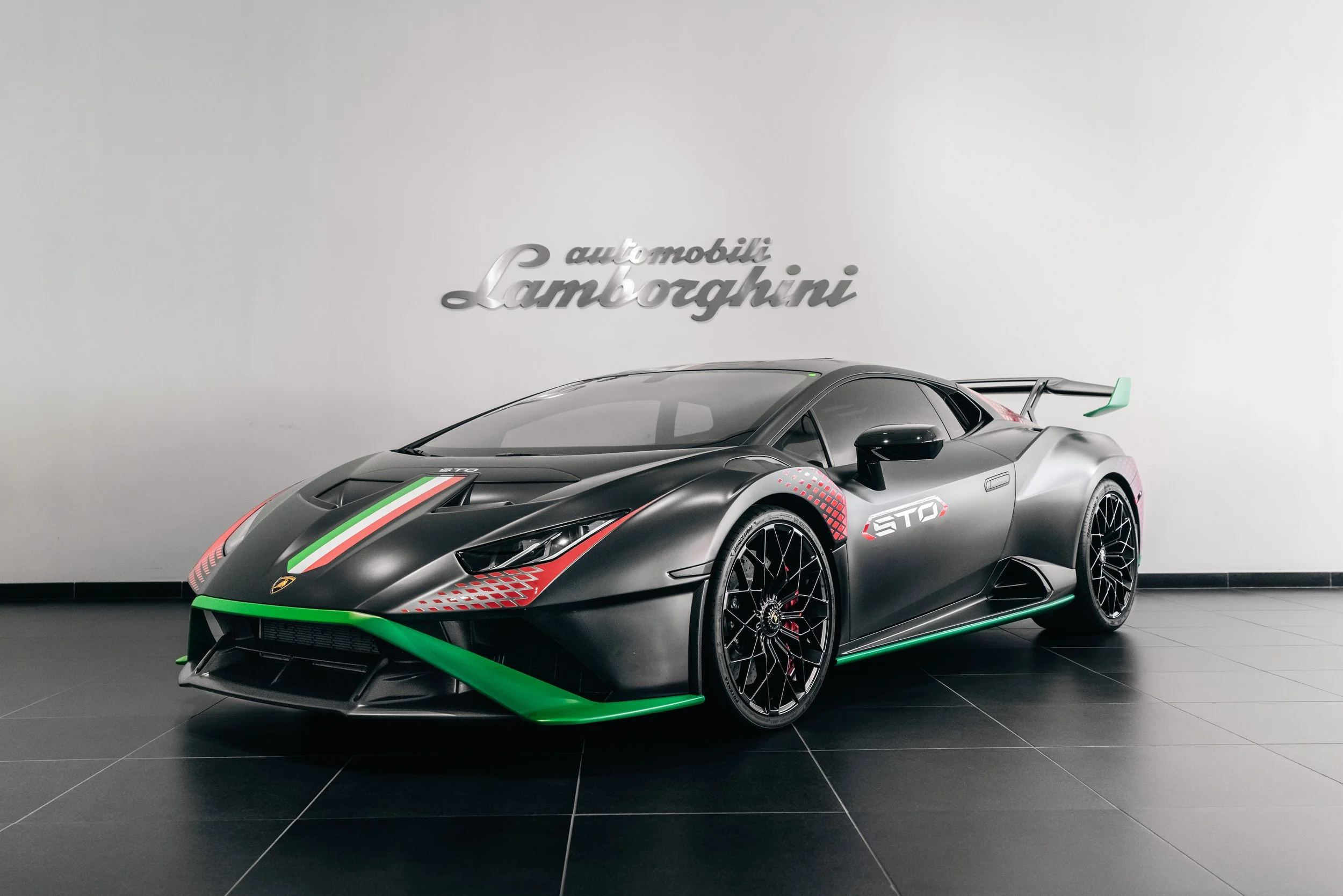 A black Lamborghini sports car with red and green racing details is displayed indoors against a white wall with a Lamborghini logo.