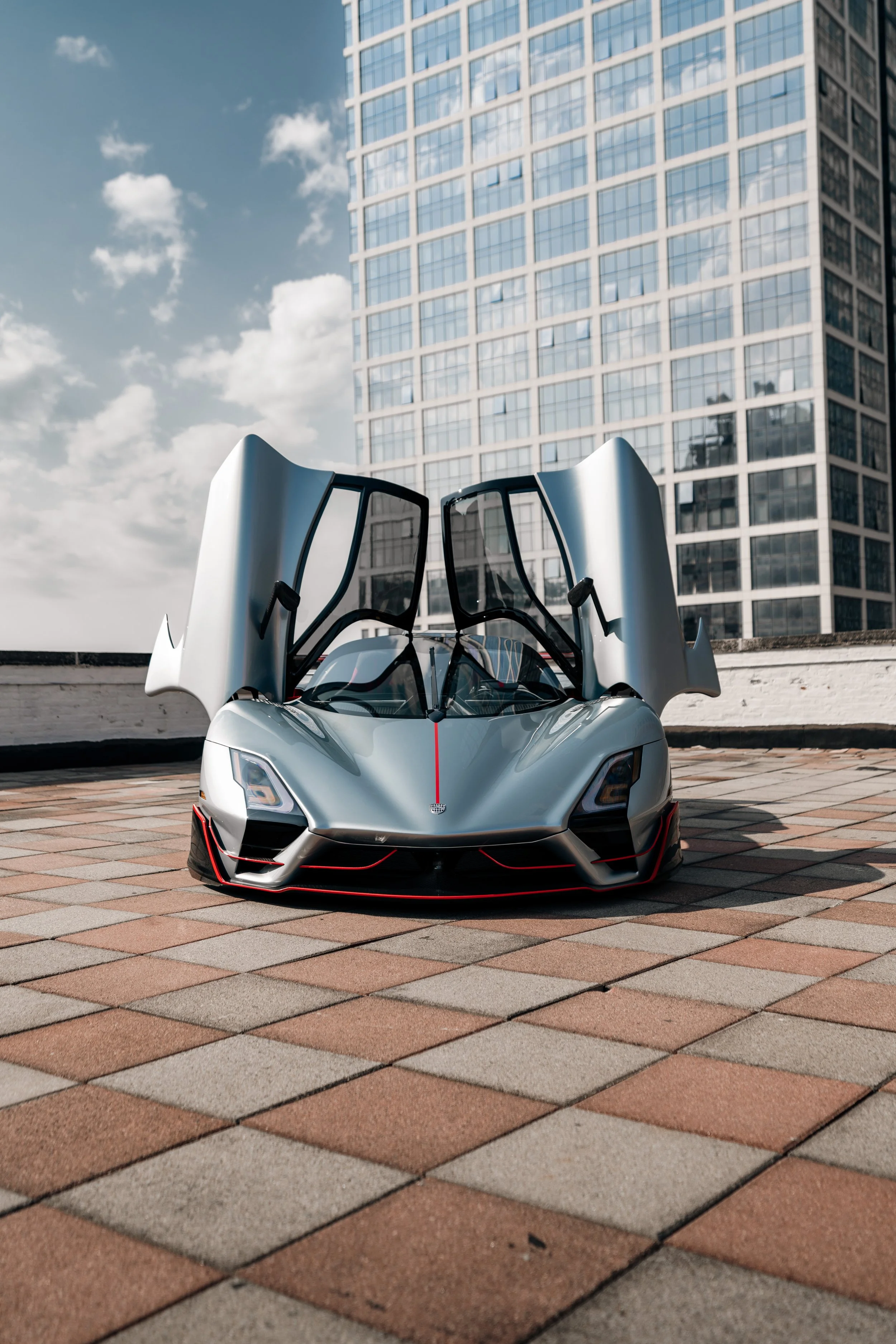 A silver sports car with its gullwing doors open, parked on a brick-paved area in an urban setting with a modern glass office building in the background.