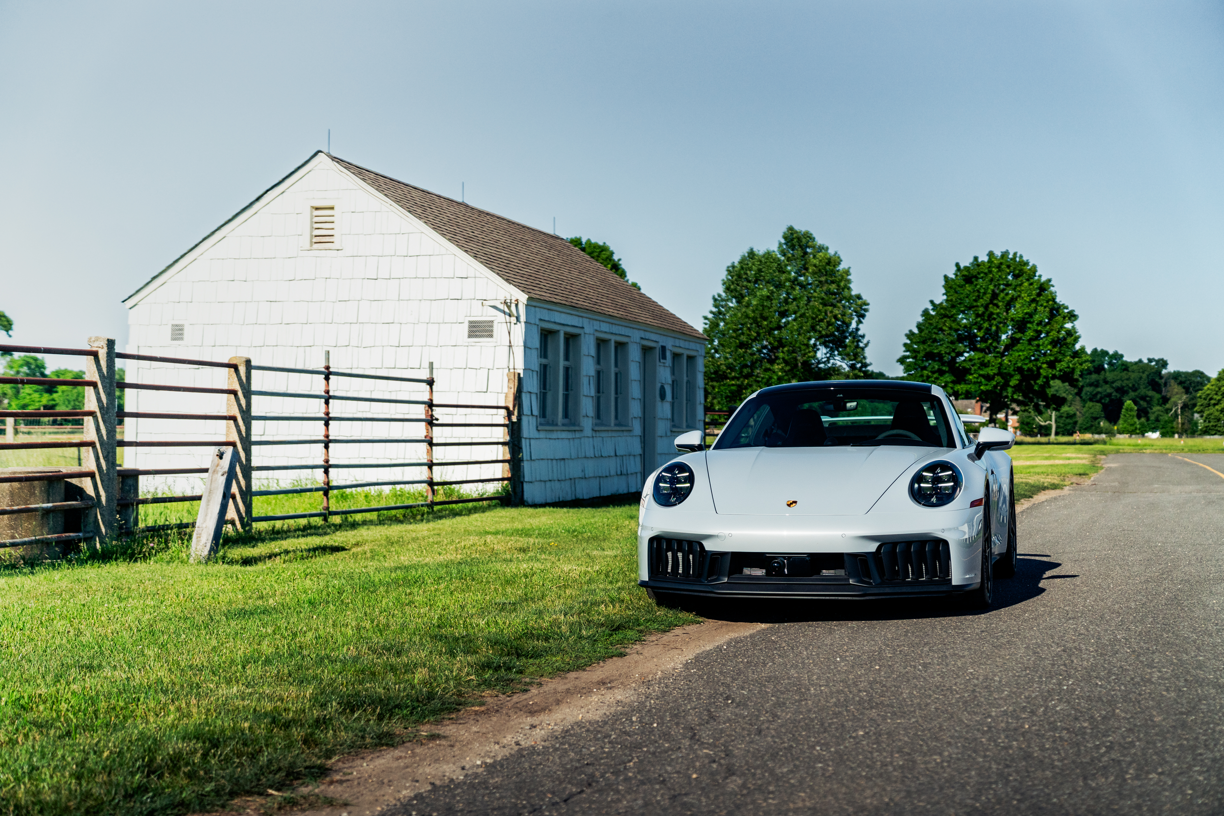 A Porsche 911 GTS T-Hybrid parked on a rural road with green grass, trees, a white shed, and a rusty fence in the background.