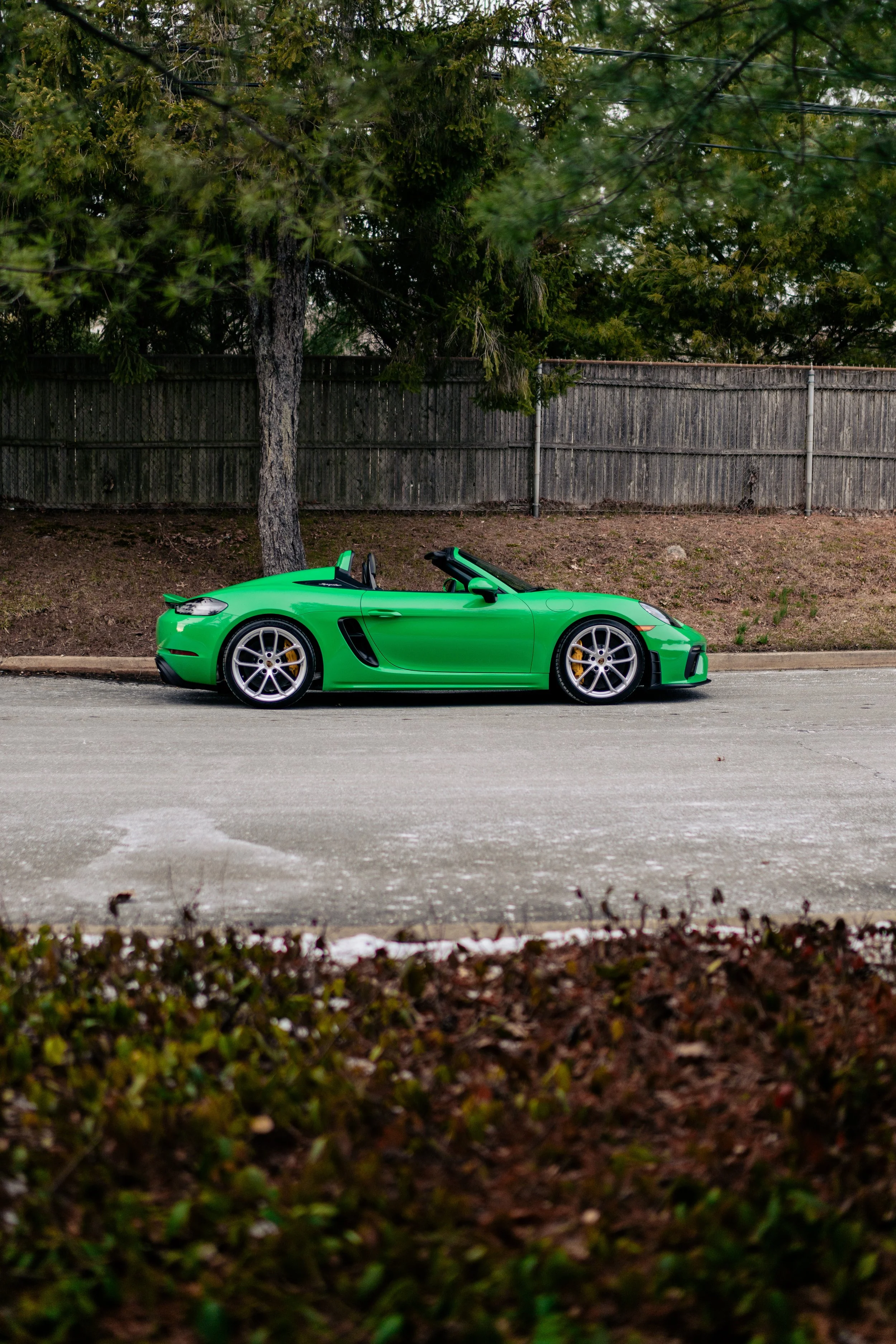A bright green sports car with its top down parked on the side of a street, with a wooden fence and trees in the background.