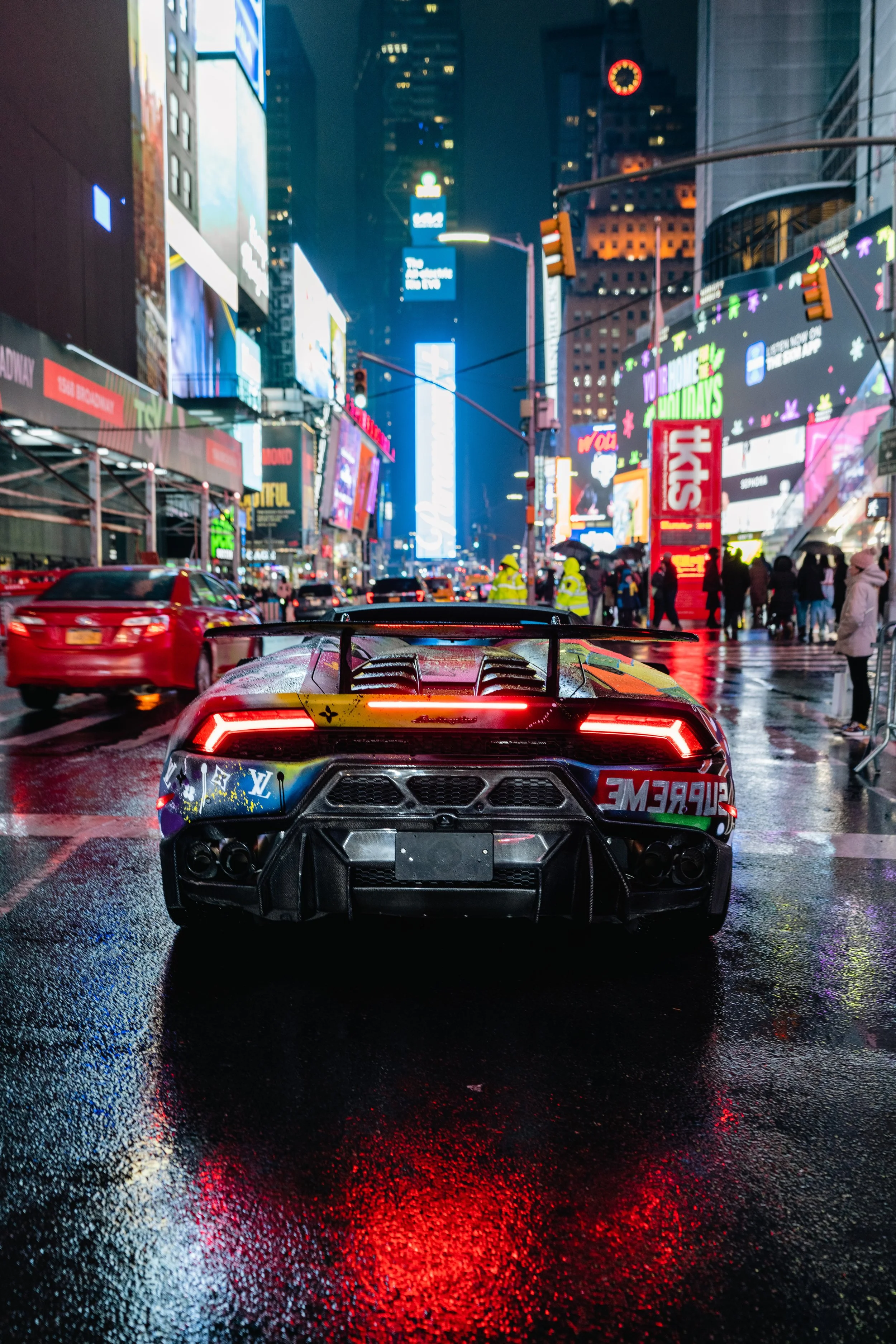 A sports car with a colorful wrap is parked on a wet city street at night, with bright neon signs and billboards in the background, and pedestrians walking by.