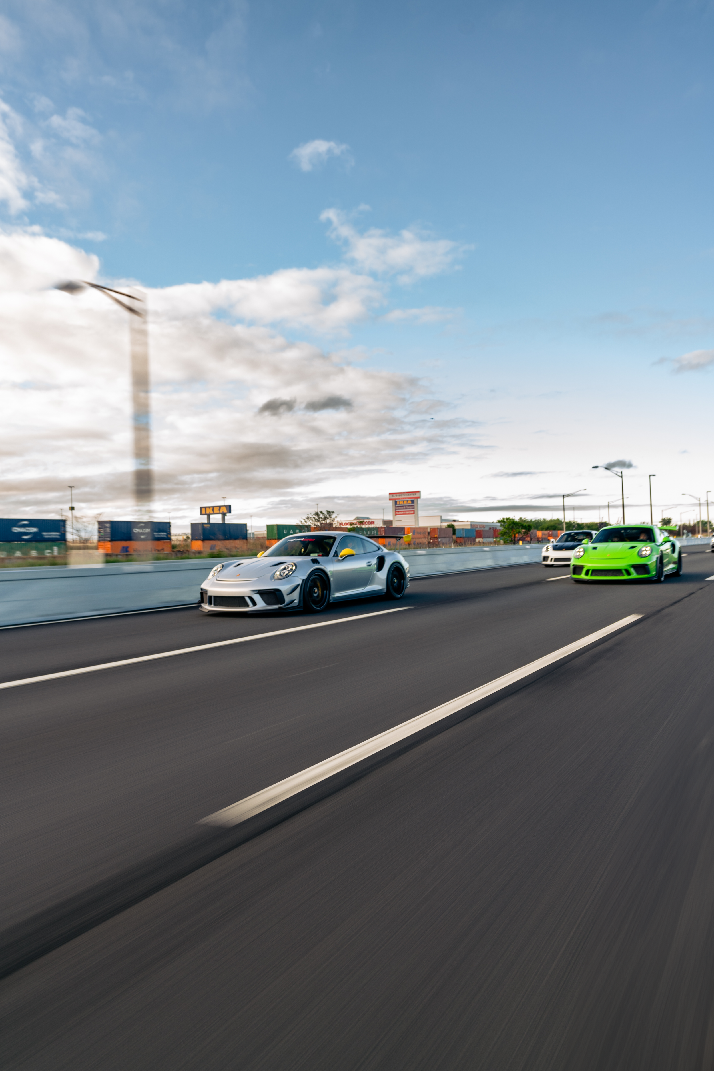 Two sports cars, one gray and one bright green, driving on a highway with a cloudy sky and shipping containers in the background.