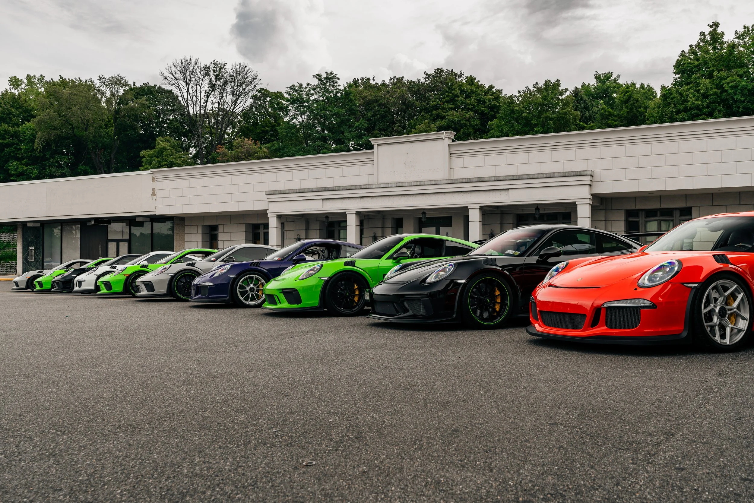 Lineup of sports cars, mainly Porsche models, parked outside a white building with trees in the background, under an cloudy sky.