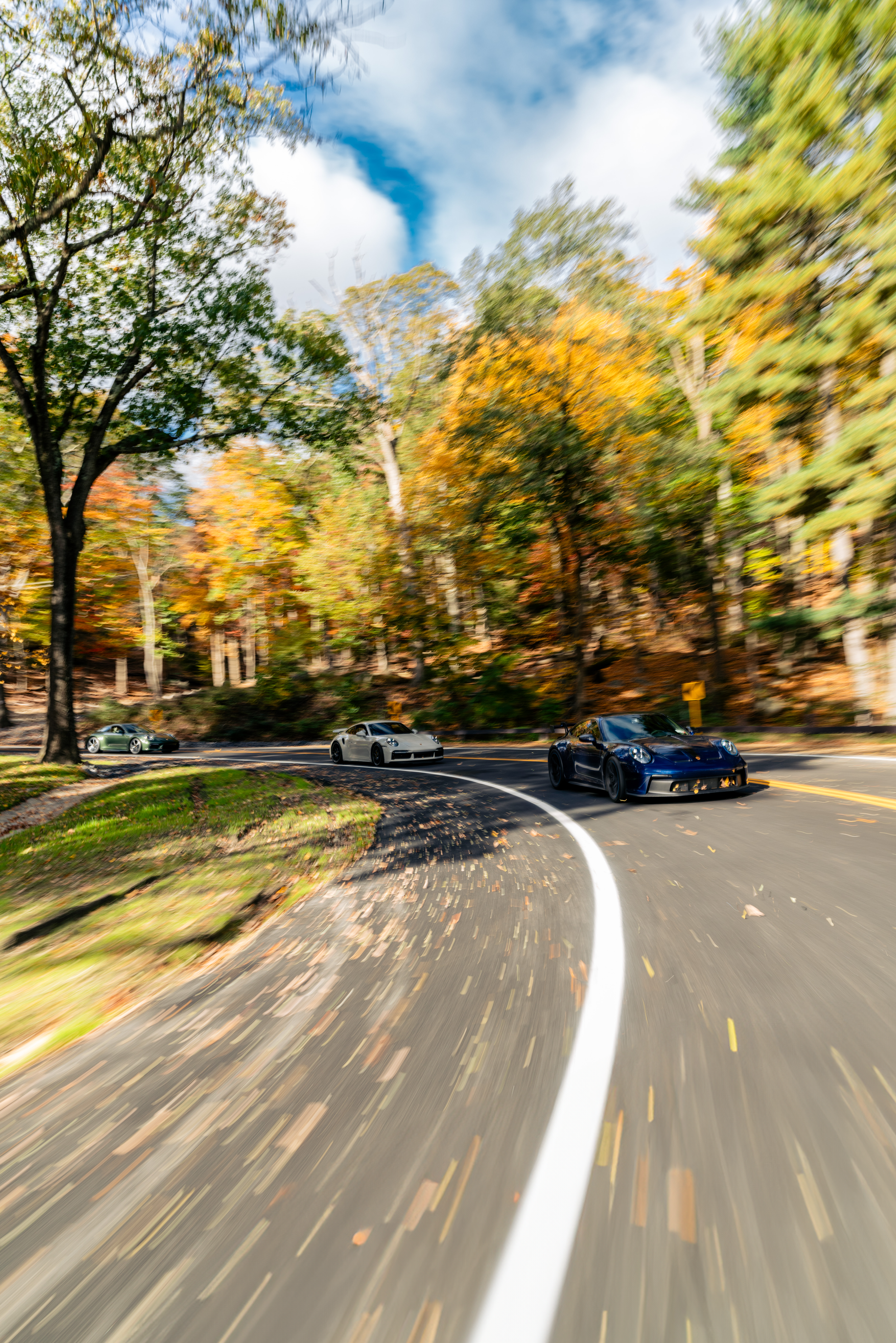 A curved road with cars driving on it, surrounded by trees with autumn-colored leaves, under a partly cloudy sky.