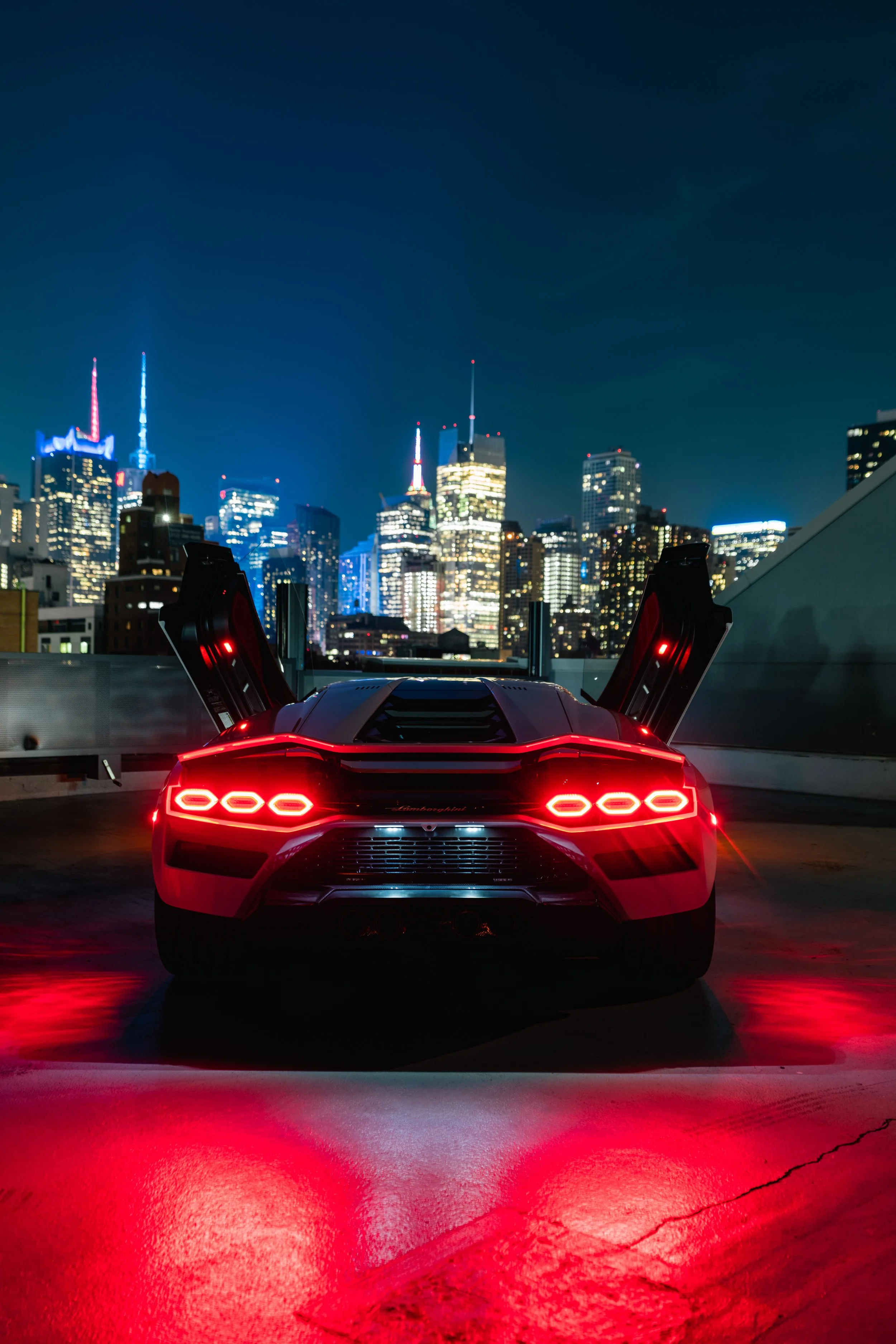 Rear view of a red Lamborghini sports car with scissor doors open, city skyline at night with tall lit-up skyscrapers in the background, and red reflections on the wet asphalt.