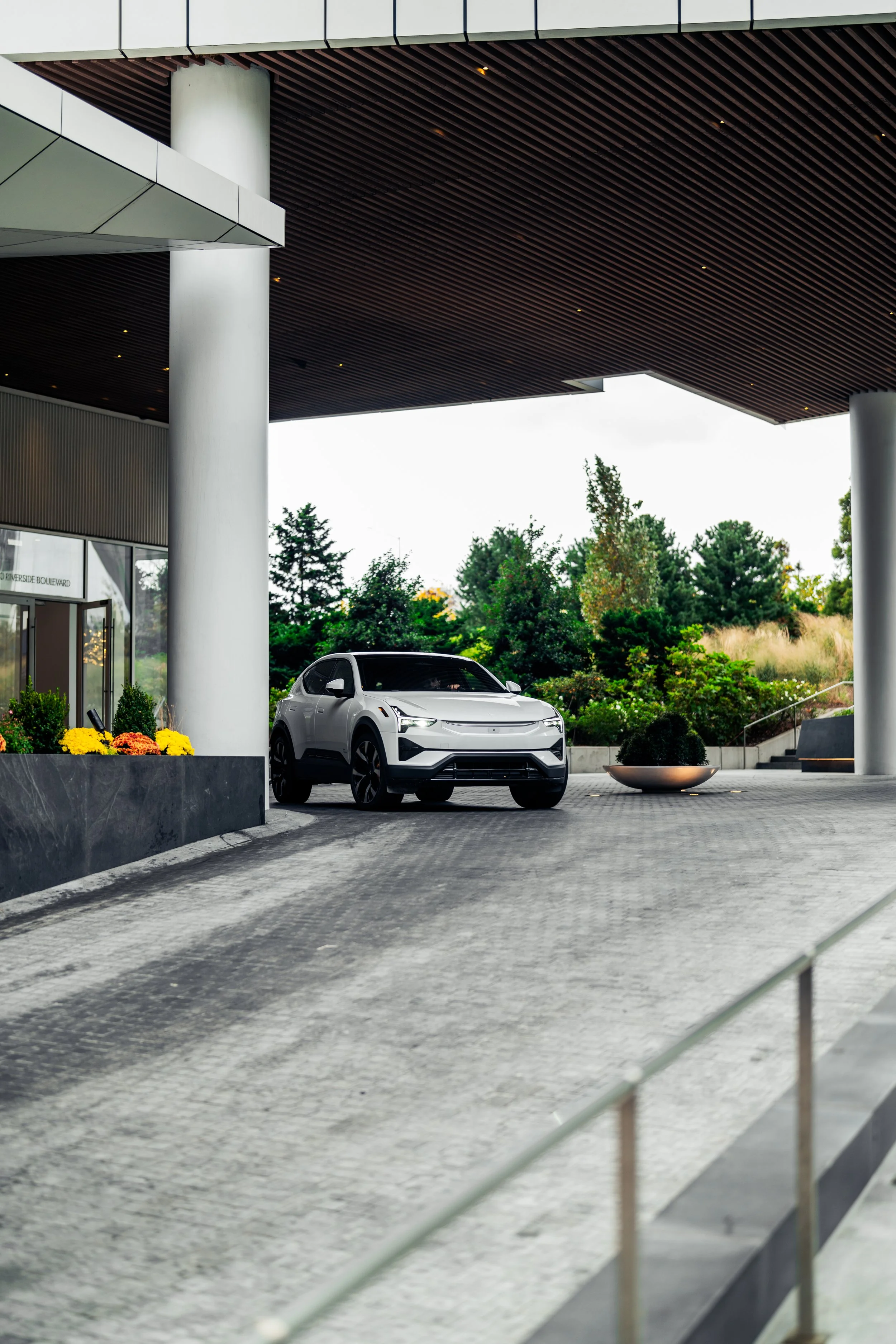 A white electric car parked under a modern building with a wooden ceiling, surrounded by greenery, with yellow flowers in a planter in the foreground.