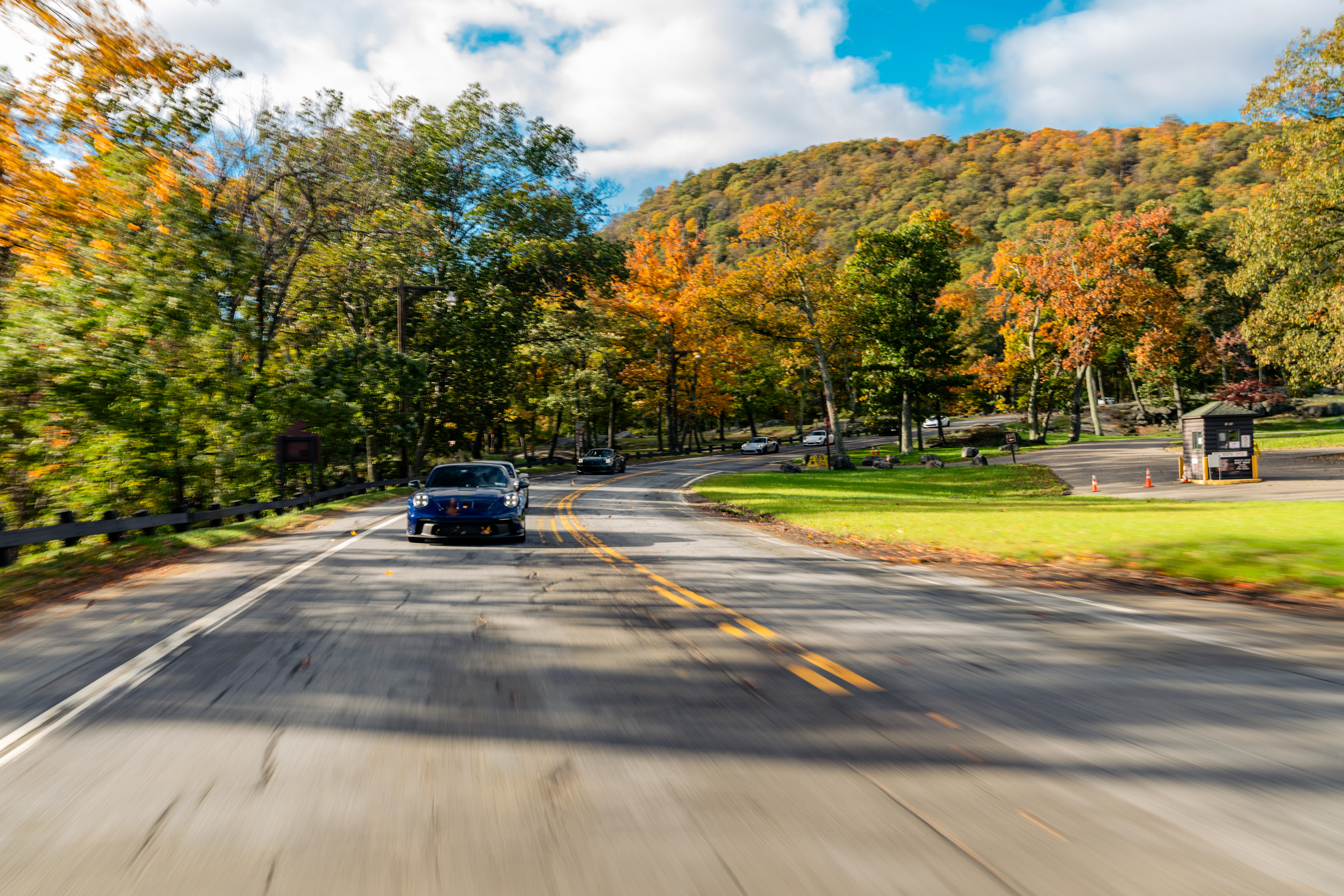 A winding road with cars passing through a forested area during autumn, with trees showing fall colors and a large hill in the background under a partly cloudy sky.