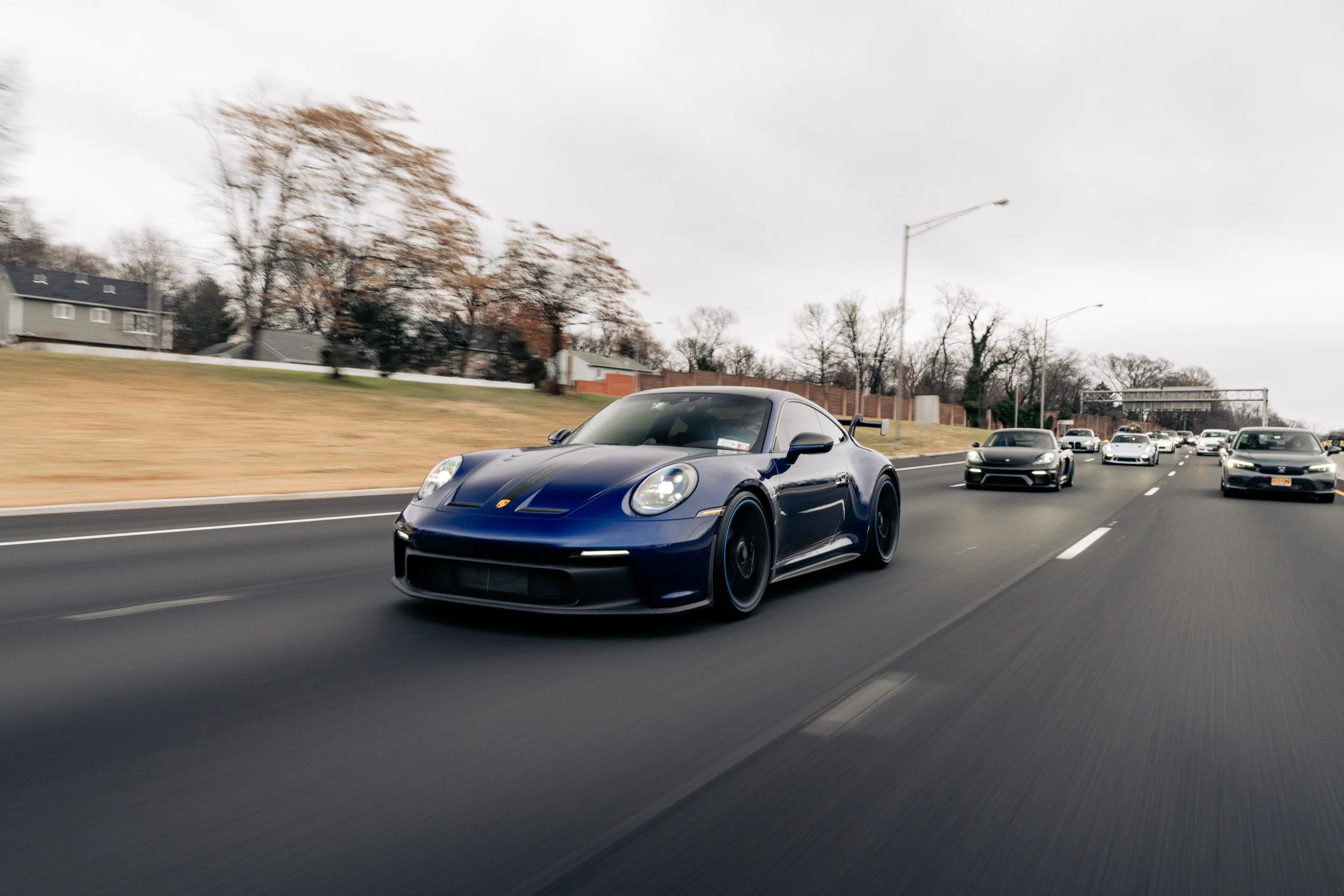 A blue sports car driving on a highway with other vehicles after a rainstorm, with trees and houses in the background, overcast sky.