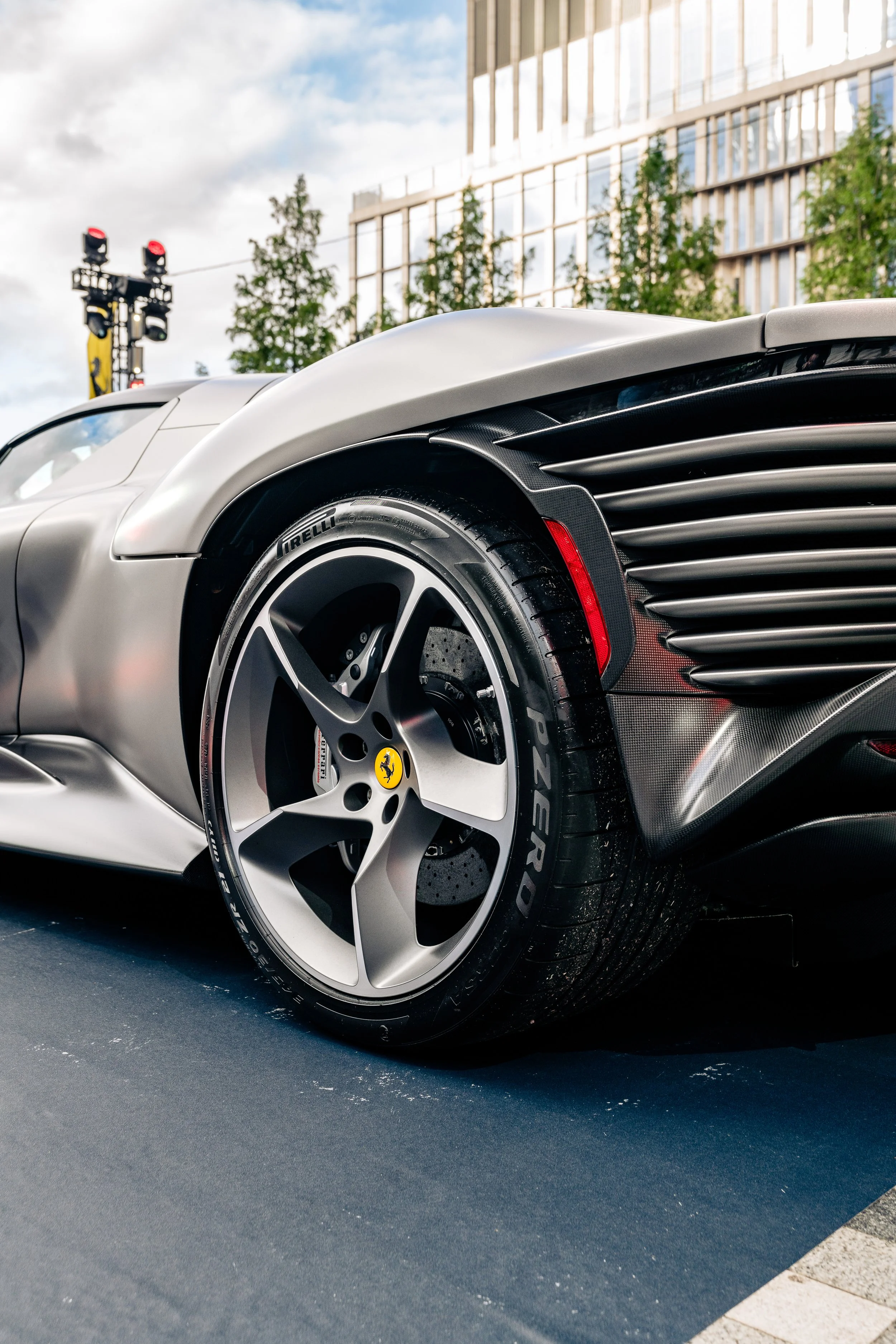 Close-up of a silver sports car with a Ferrari badge on the wheel, featuring sleek design and black accents, parked in an outdoor setting near a modern glass building and green trees.