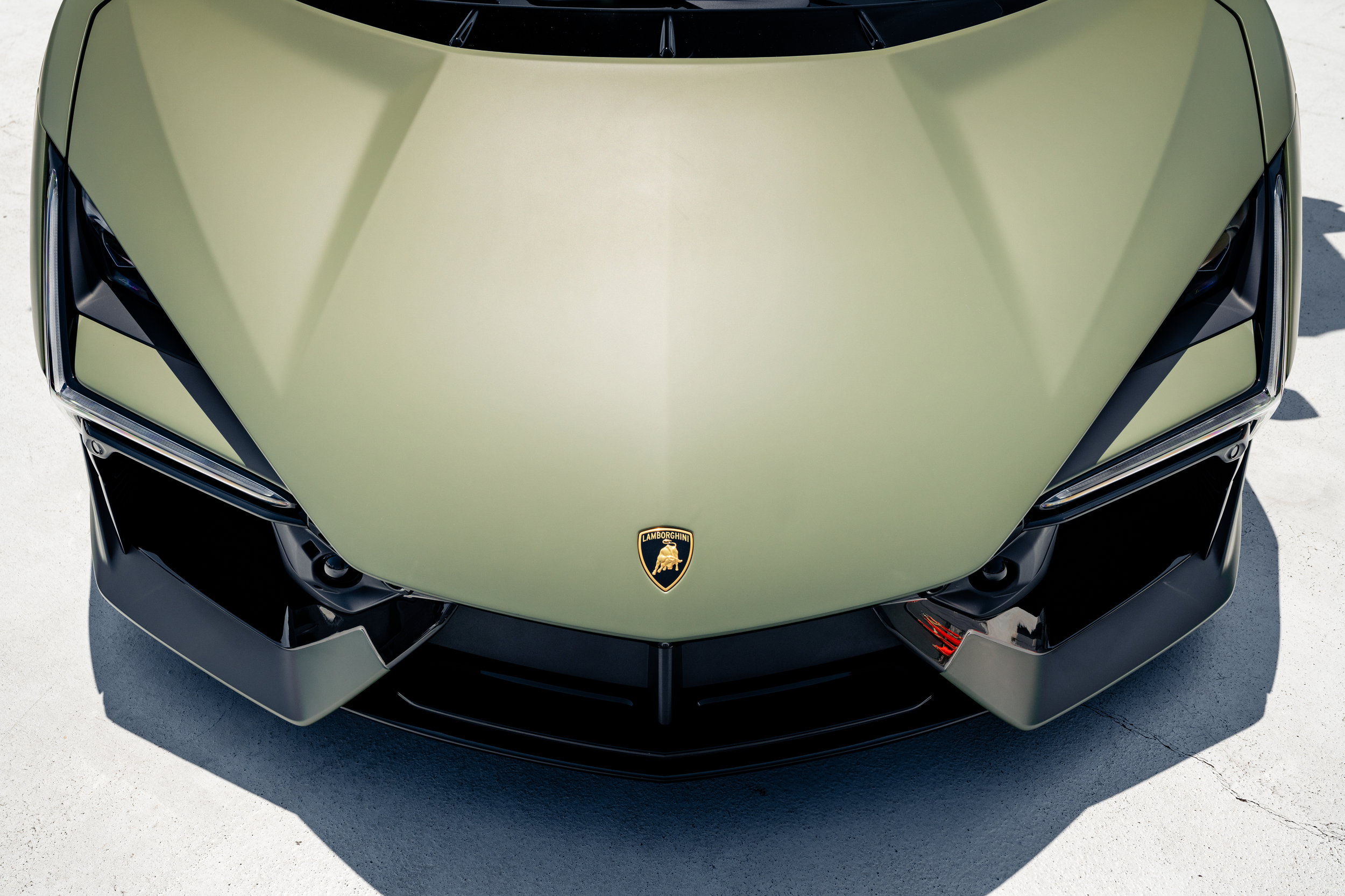 An overhead shot of a Lamborghini Revuelto with a matte green hood and black details, parked on a light-colored concrete surface.