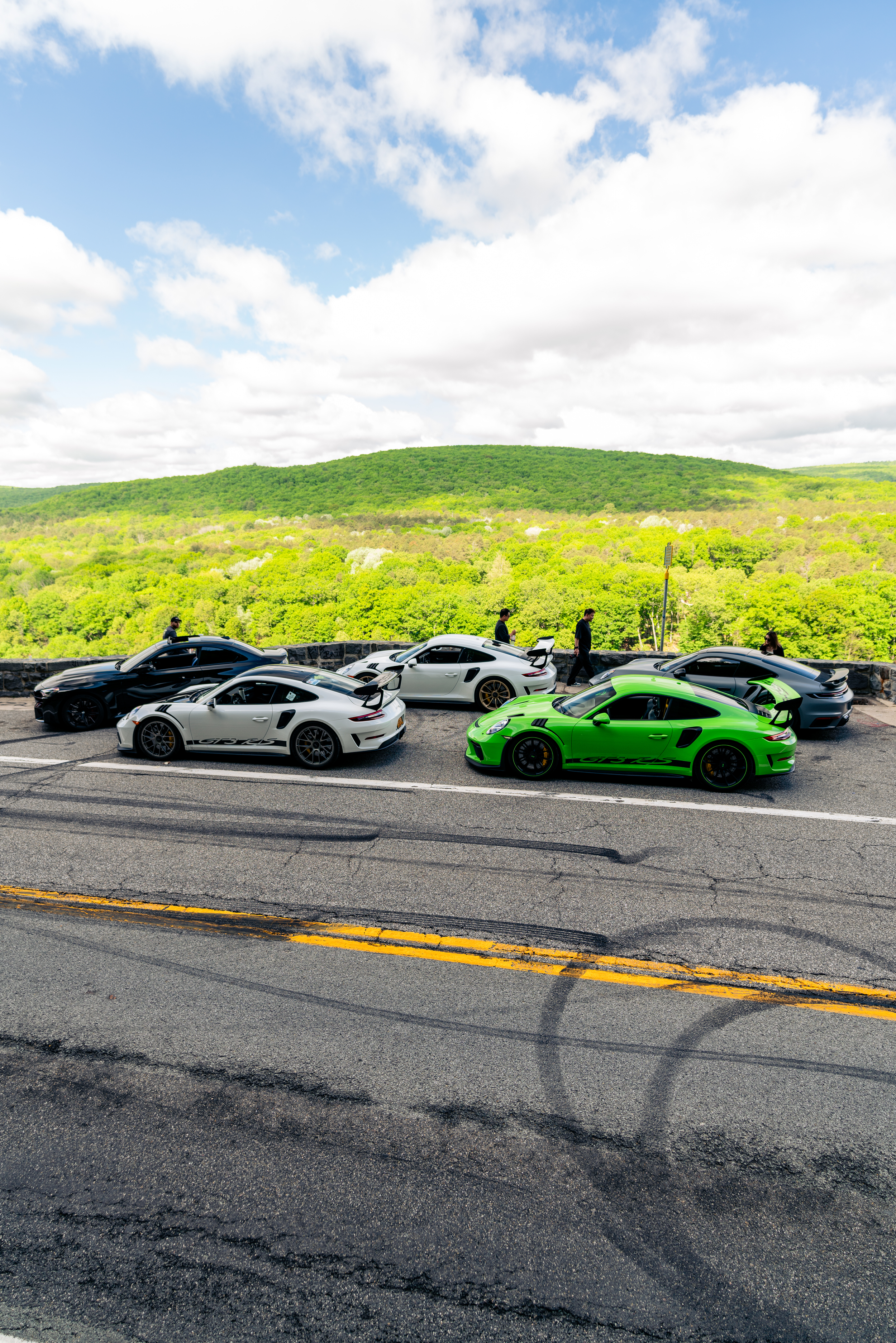 Five sports cars, including a green Porsche and a white Porsche, parked along a street with a green hill and cloudy sky in the background, and a few people walking nearby.
