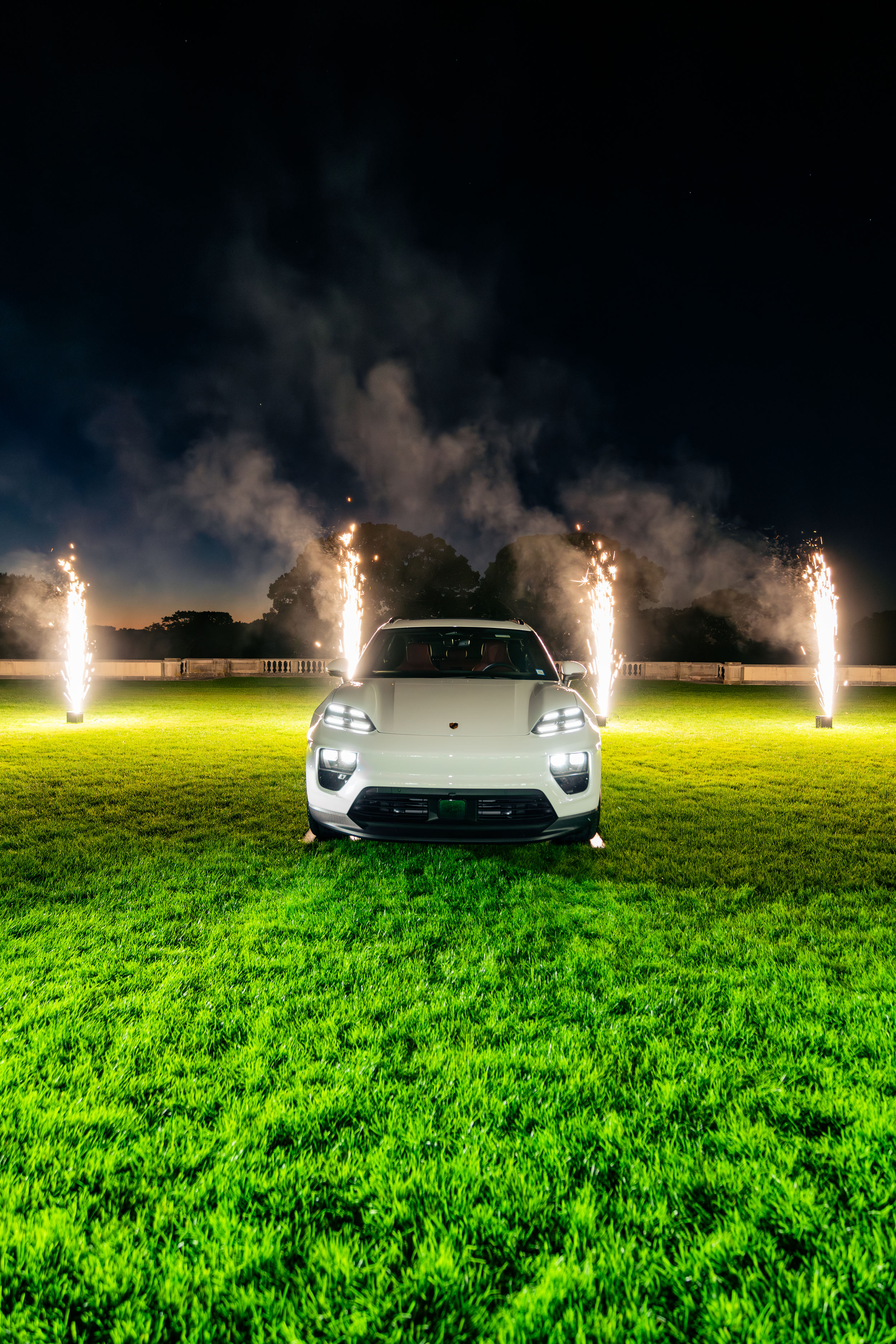A white sports car with bright headlights parked on a lush green lawn at night with fireworks and smoke in the background.